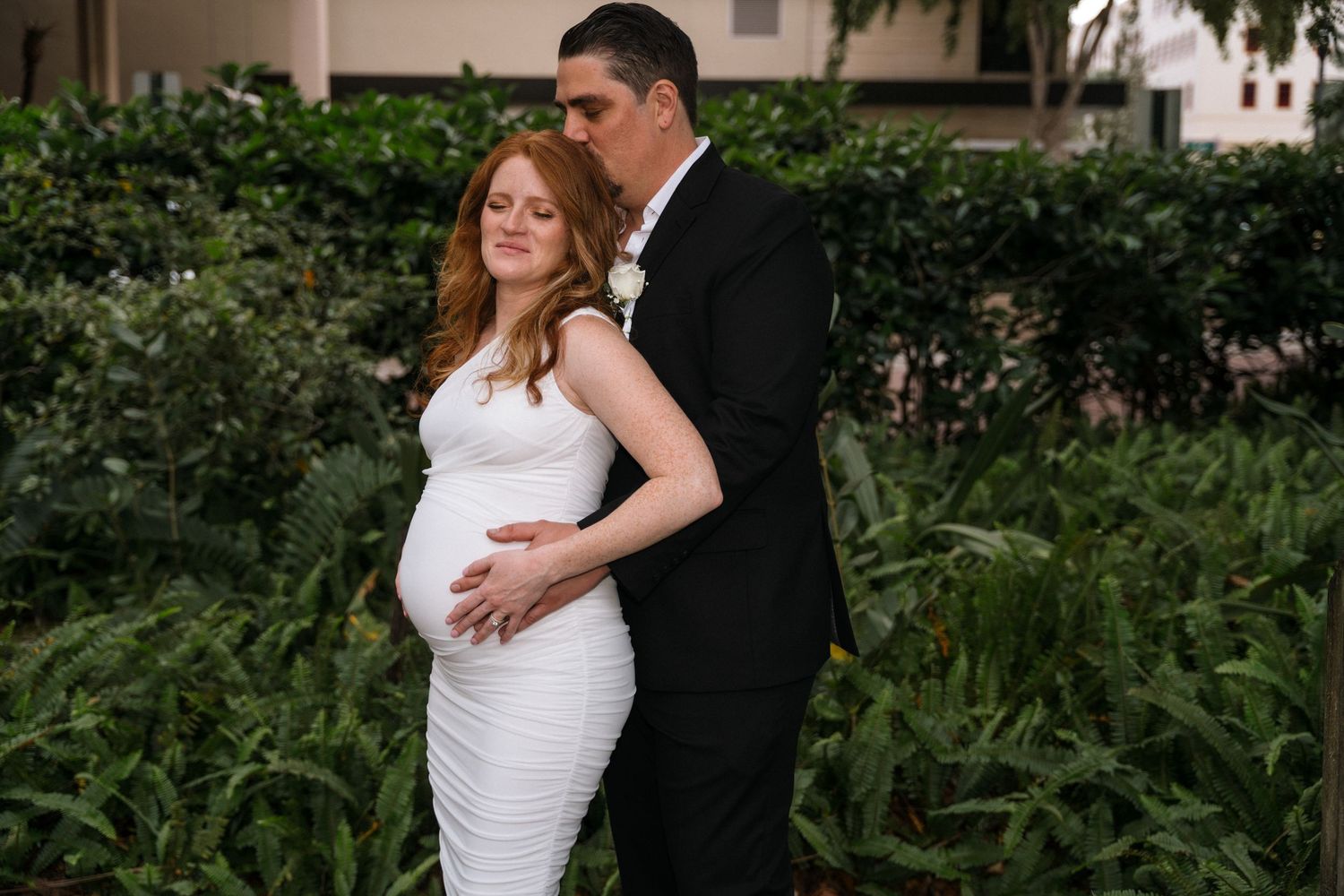 Romantic maternity wedding photo of couple embracing in a garden setting.