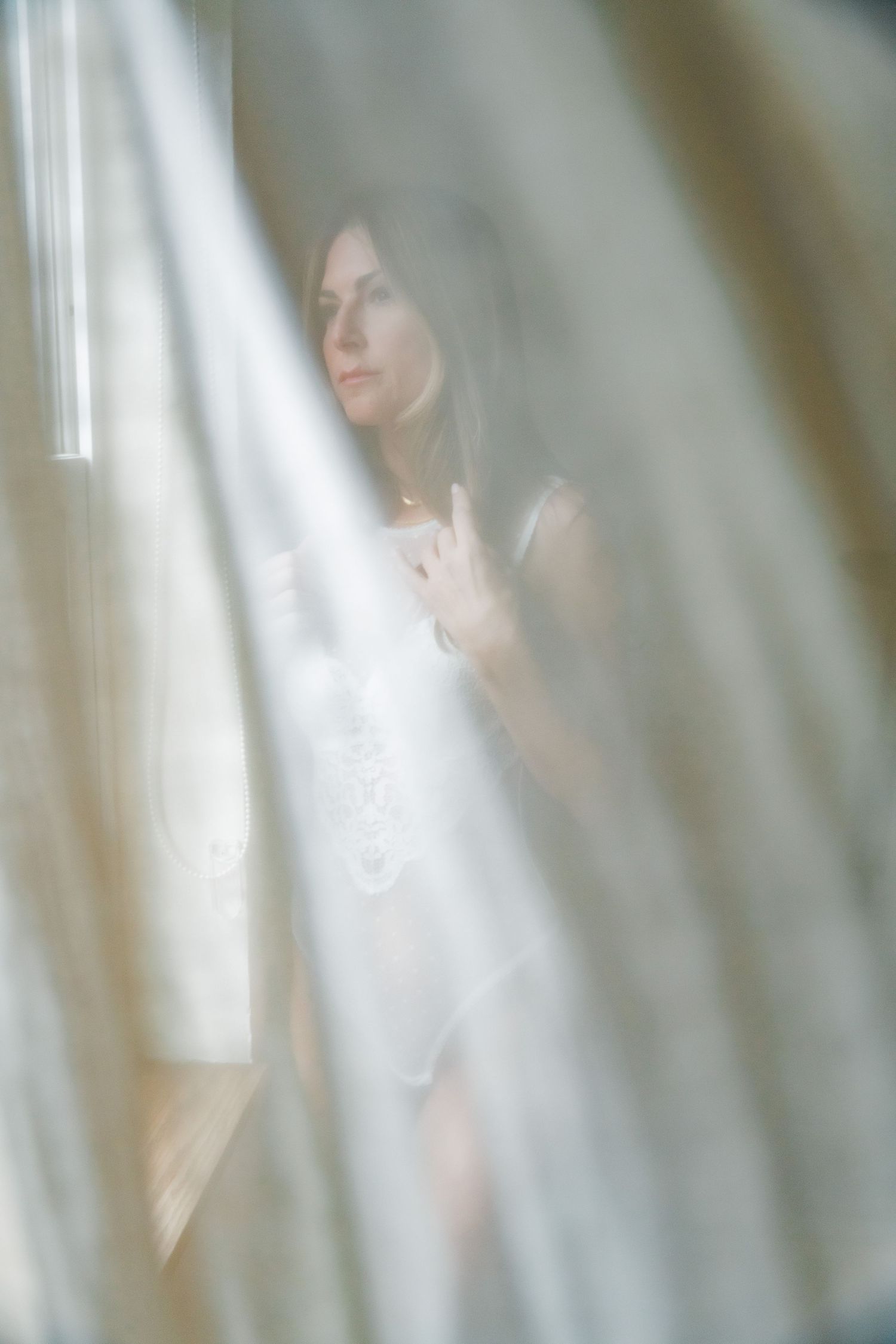 Ethereal portrait through sheer white curtains creates dreamy atmosphere with soft natural window light.