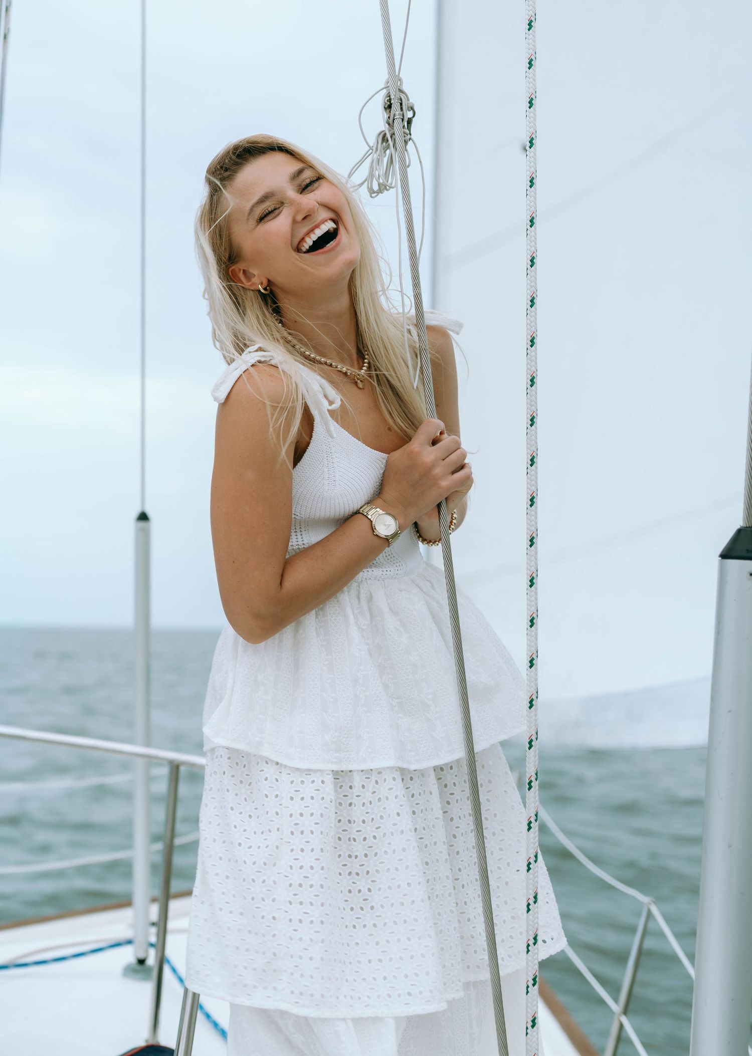 Laughing blonde woman in white dress holding sailboat mast on open water.