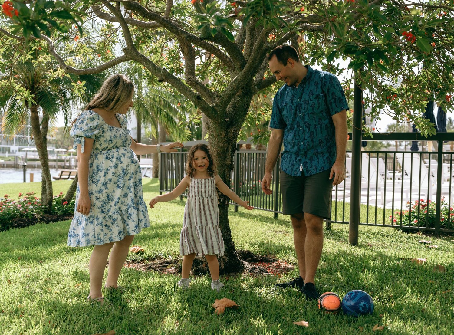 Family of three holding hands under a shady tree on green grass near a waterfront park.