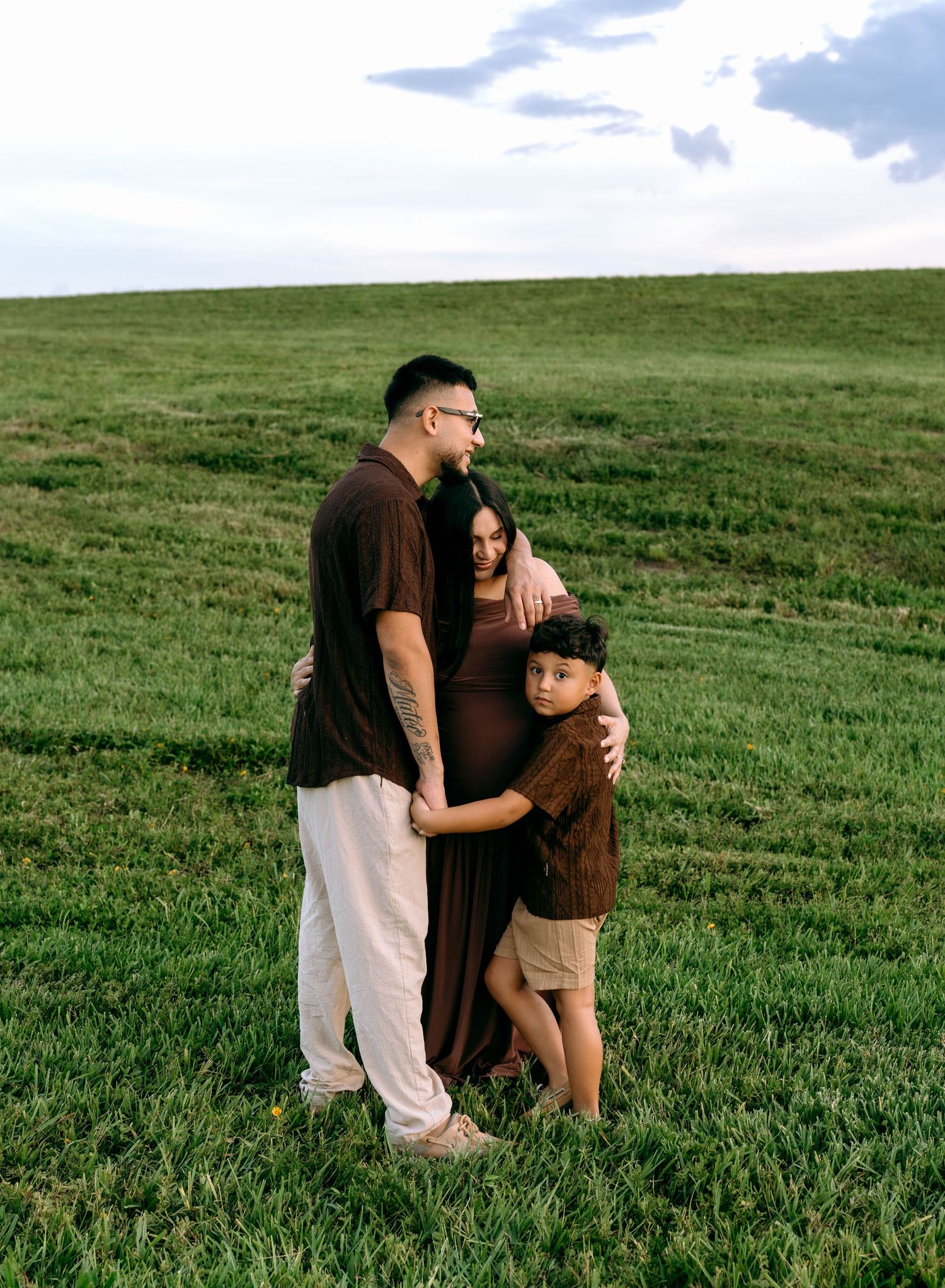 A pregnant woman in a brown dress poses with her partner and young son in a lush green field at dusk.