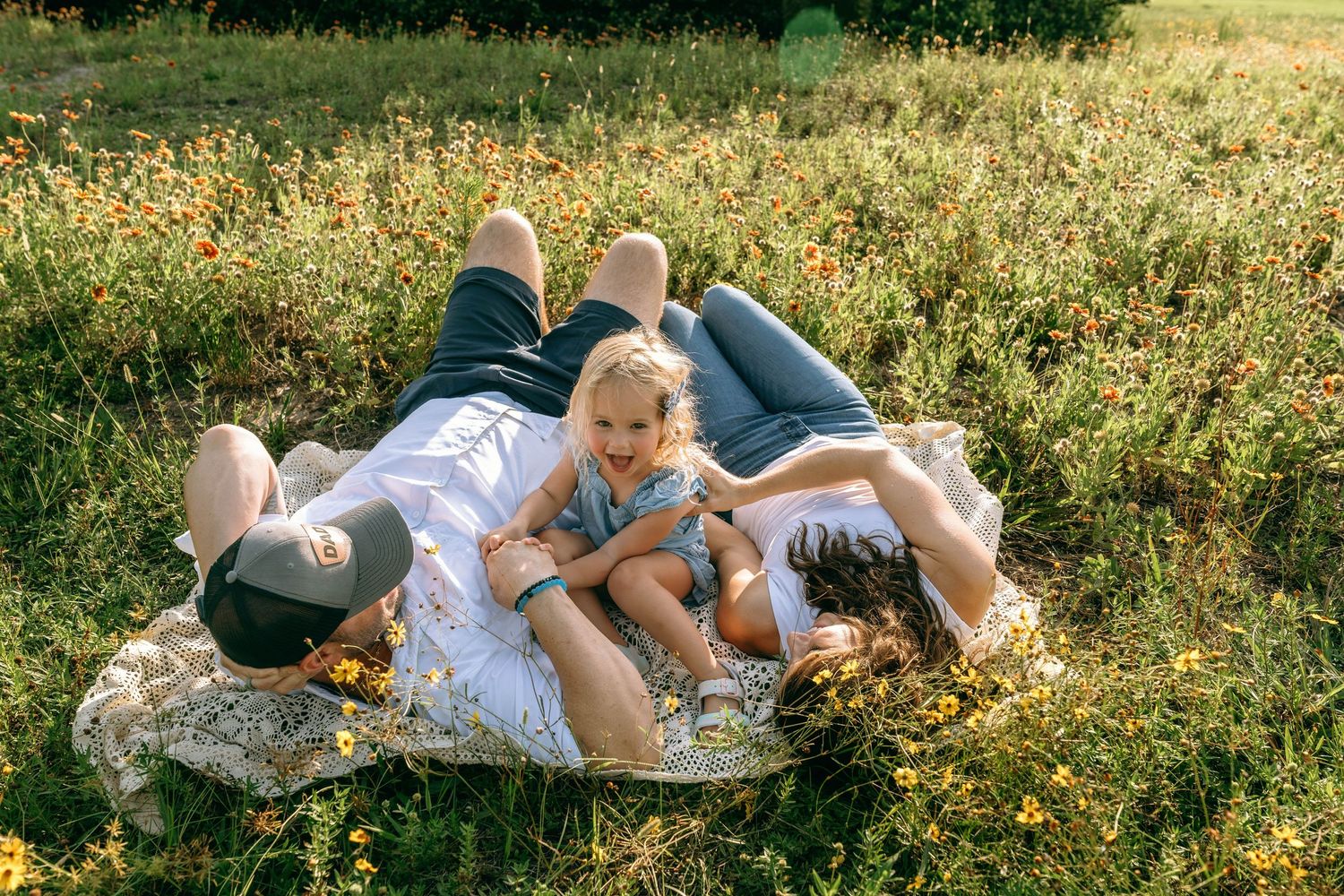 Family of three lying on a blanket in a sunny wildflower field, parents and child sharing a joyful moment outdoors.