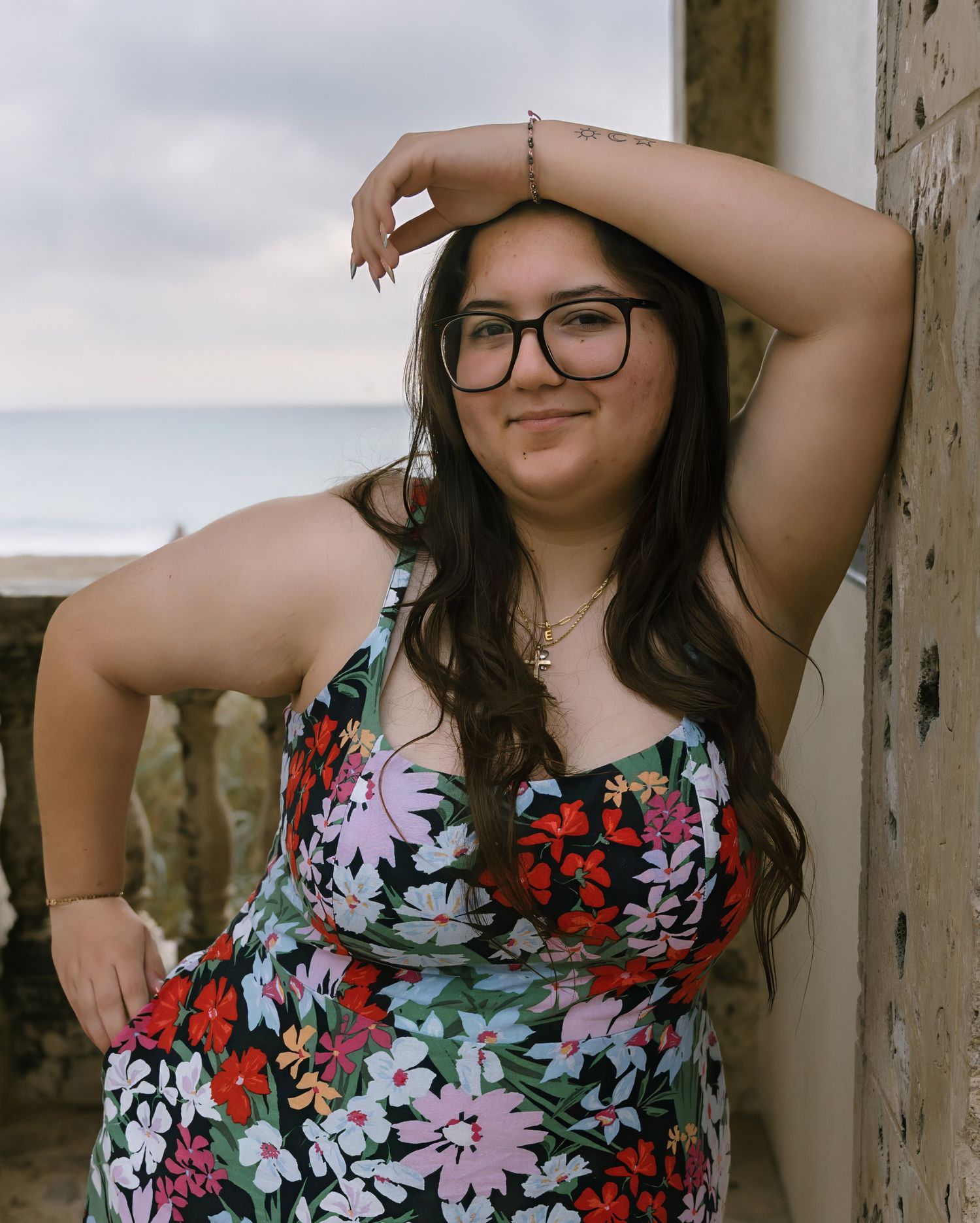 A person in glasses and a floral dress poses against a cloudy sky background.