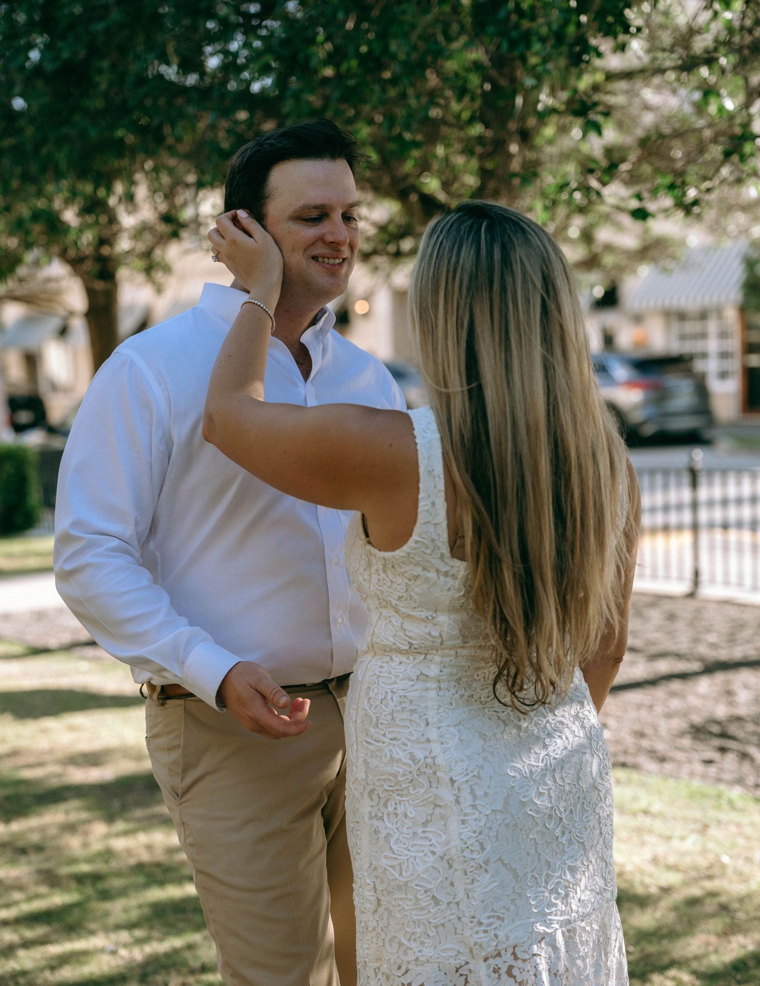 Bride in white lace dress touching grooms face as he smiles outdoors in sunlight.