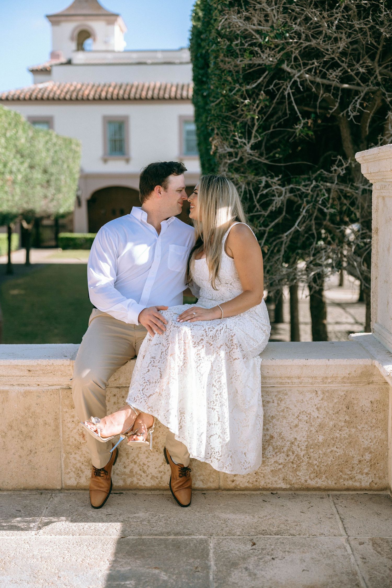 Couple sharing a romantic kiss while sitting on a stone wall outside an elegant Mediterranean-style building.