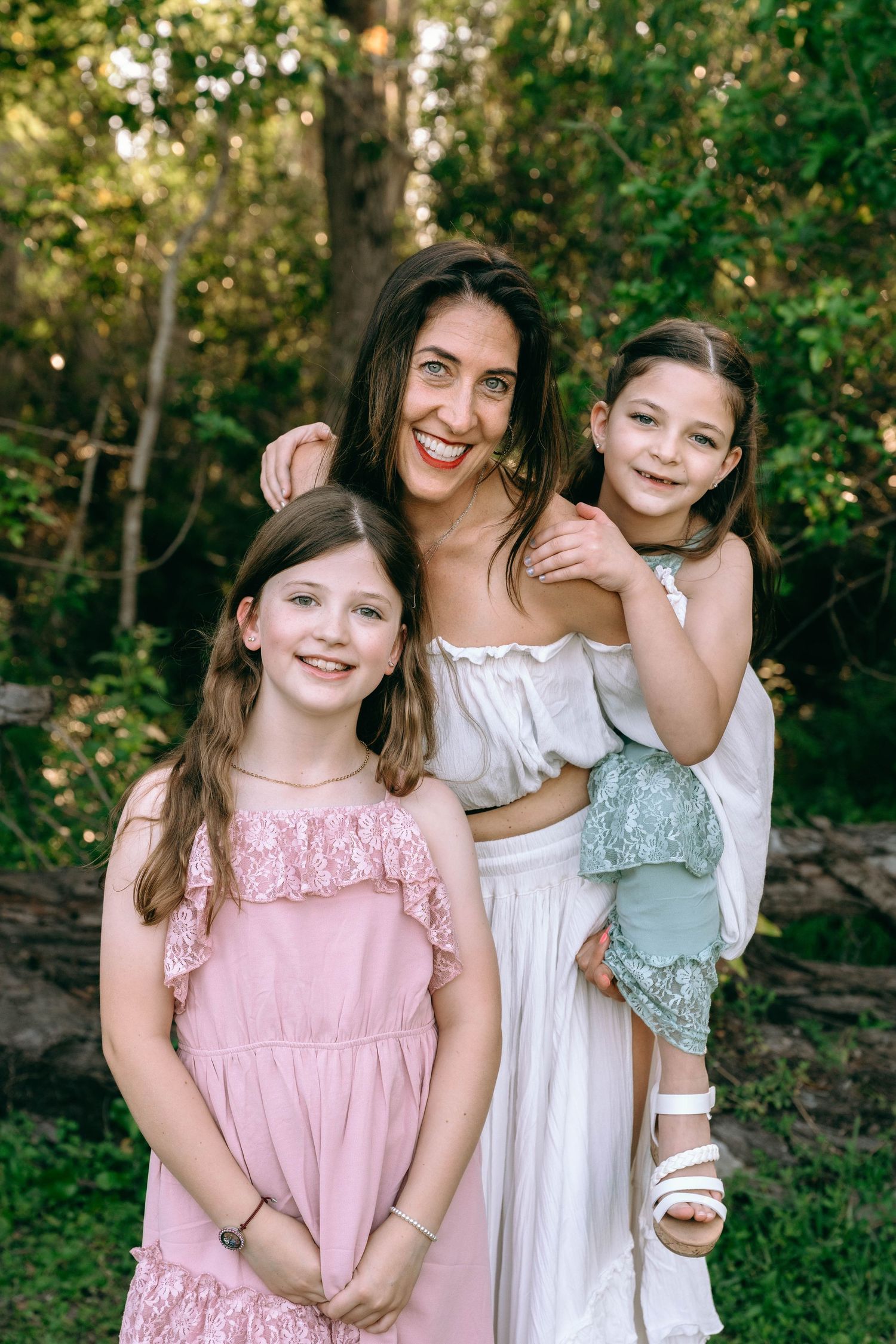 A smiling woman in white dress poses outdoors with two young girls in pink and white dresses among green trees. Colt Farms