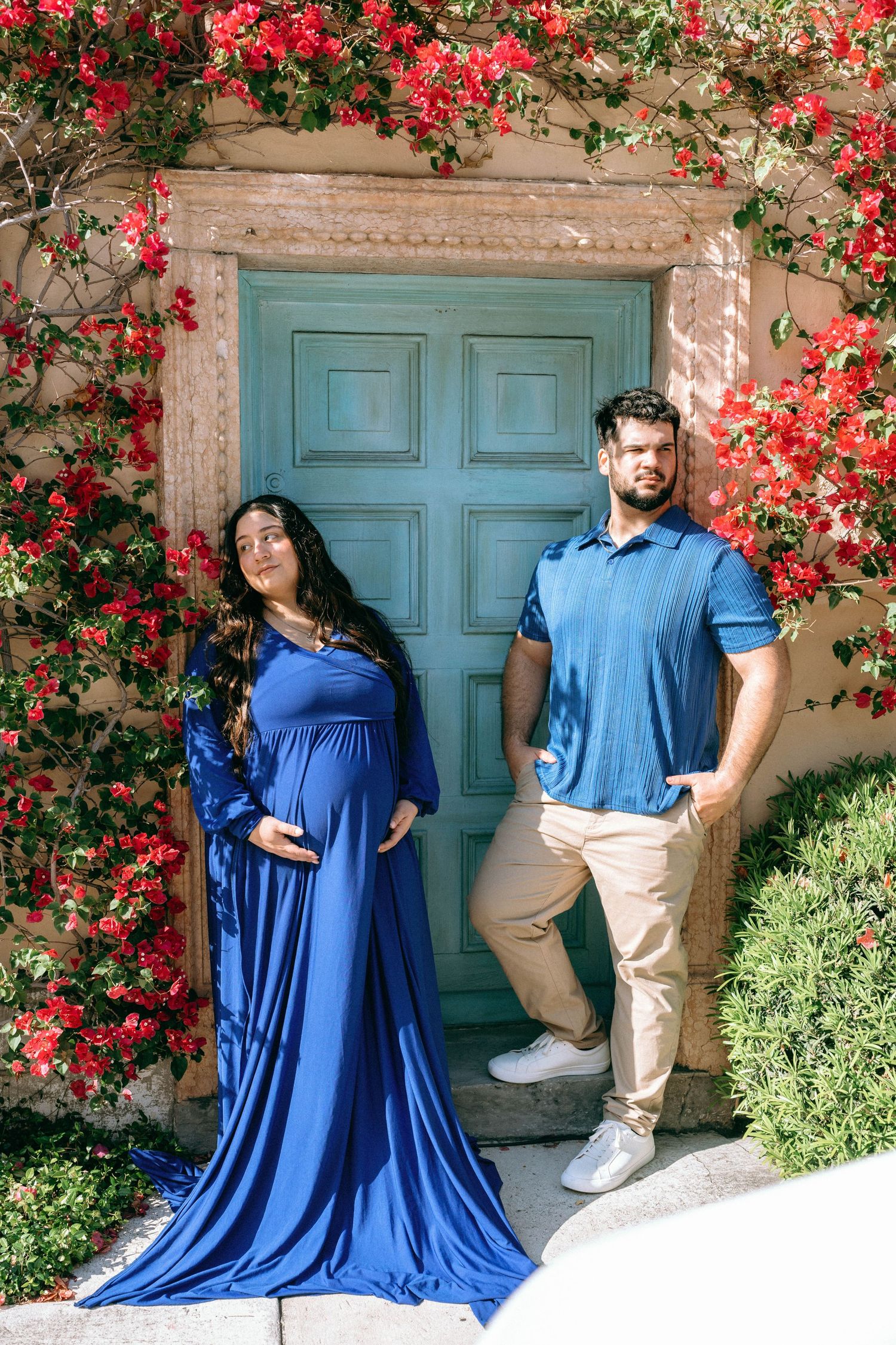 Pregnant couple posing in blue outfits before a teal door surrounded by vibrant red bougainvillea flowers. Worth Avenue, Palm Beach, FL