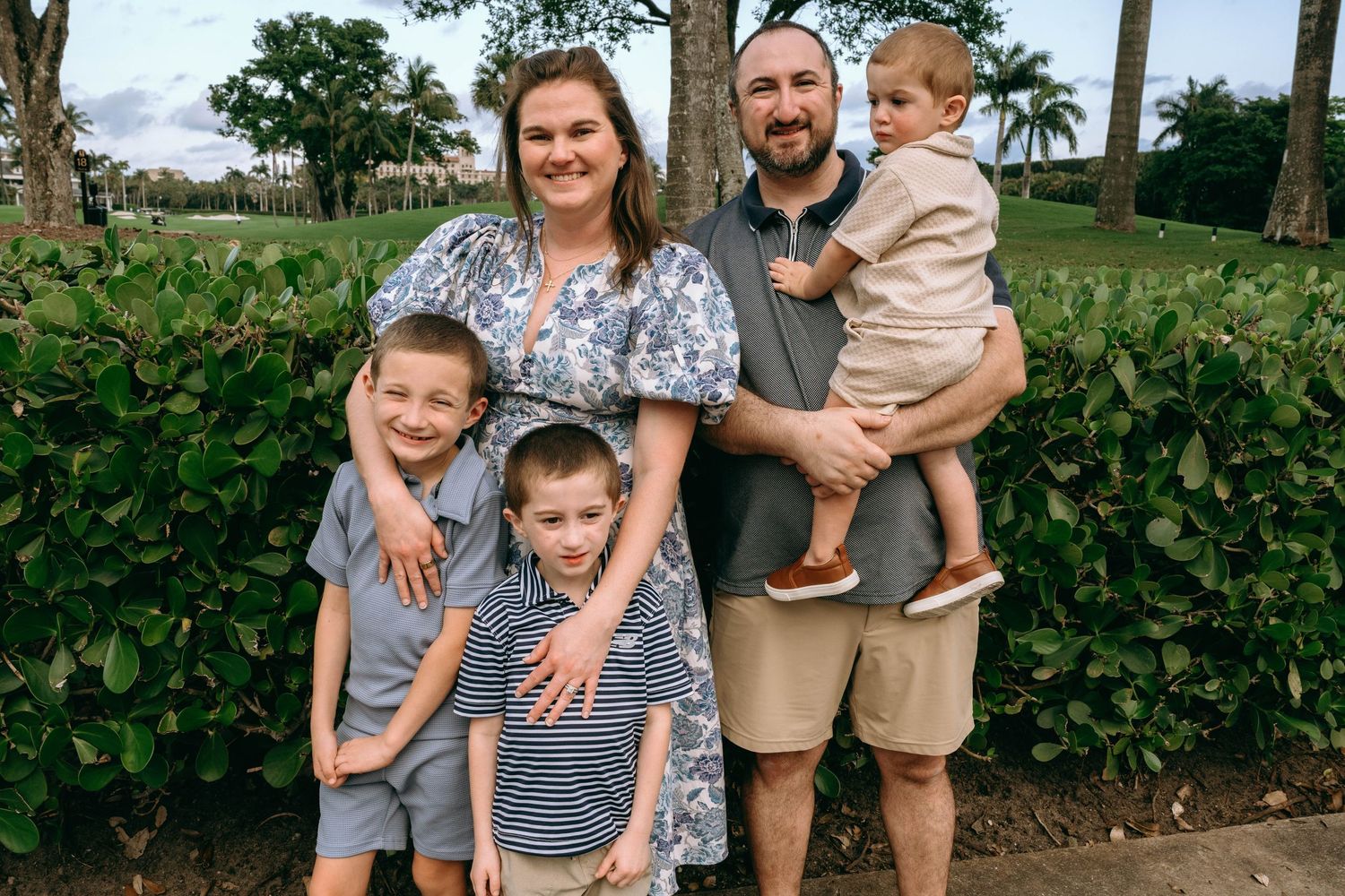 Family of five posing outdoors near lush green hedges and palm trees in a tropical setting.  Family photos at Bradley Park Palm Beach, FL