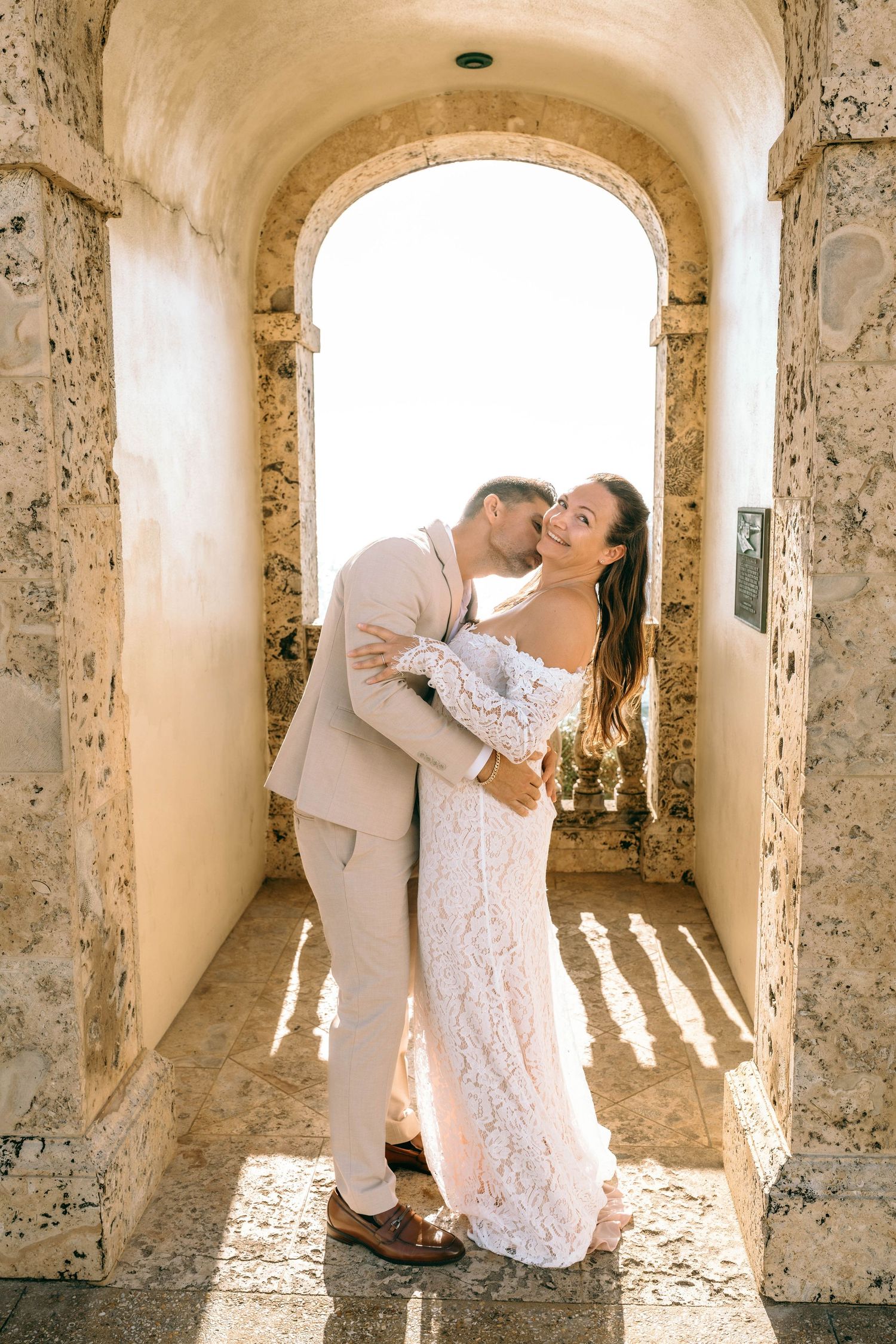Bride and groom share a romantic kiss under a stone archway in a sun-drenched corridor.