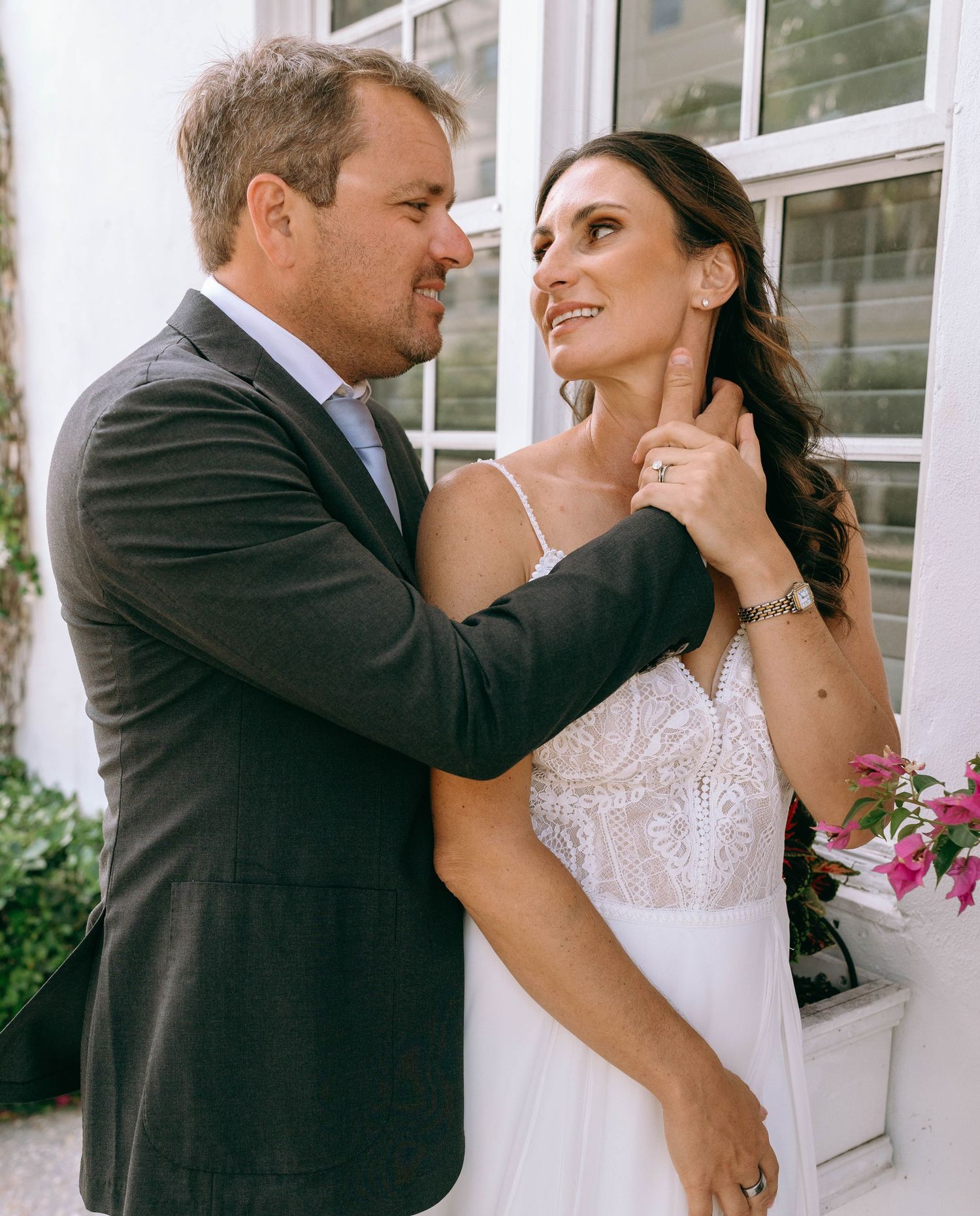 Groom tenderly adjusting brides hair near her neck while she smiles in elegant lace wedding dress.