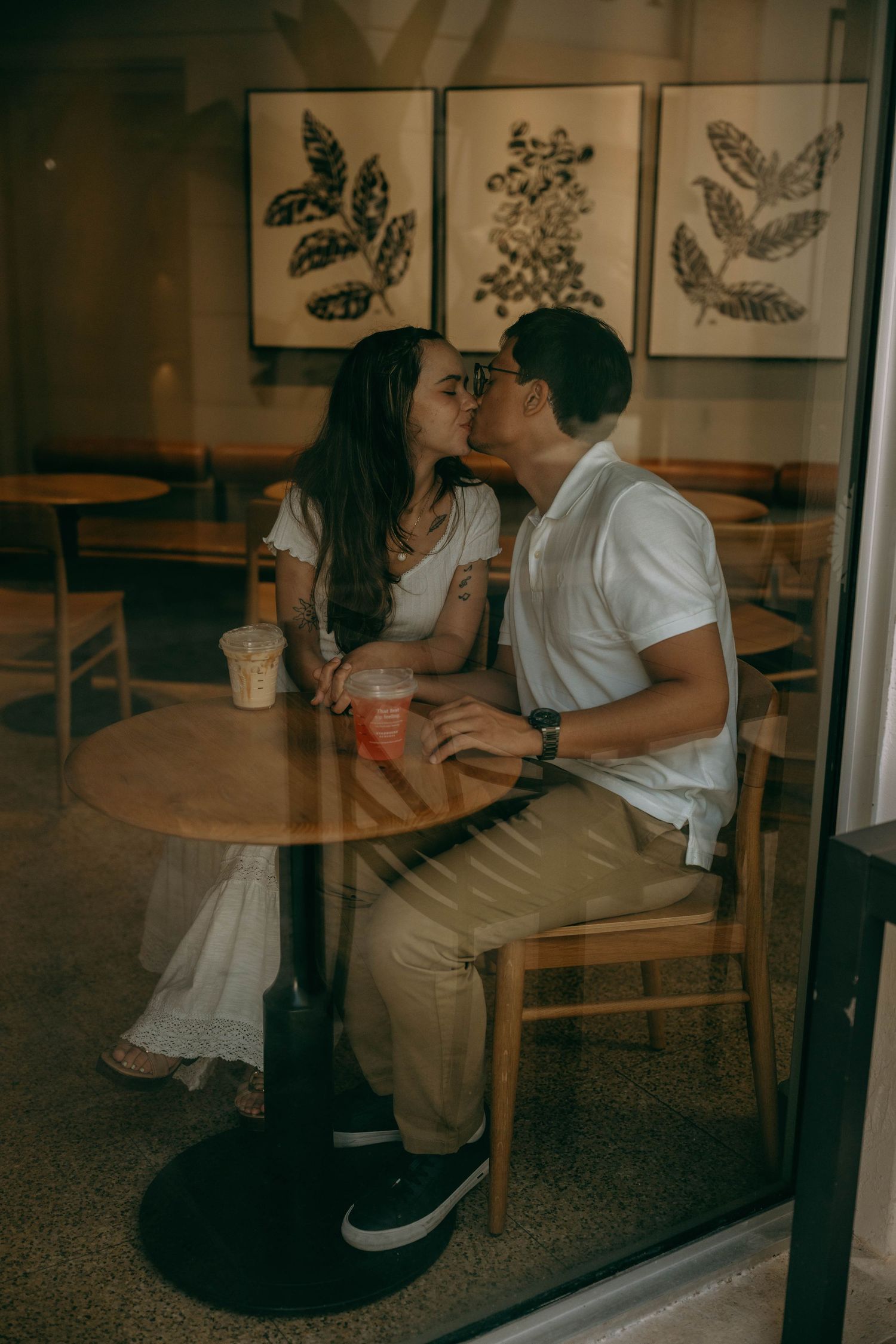 Couple sharing a kiss at a cafe table with iced drinks, botanical art prints on the wall behind them. Engagement photos on Worth Avenue