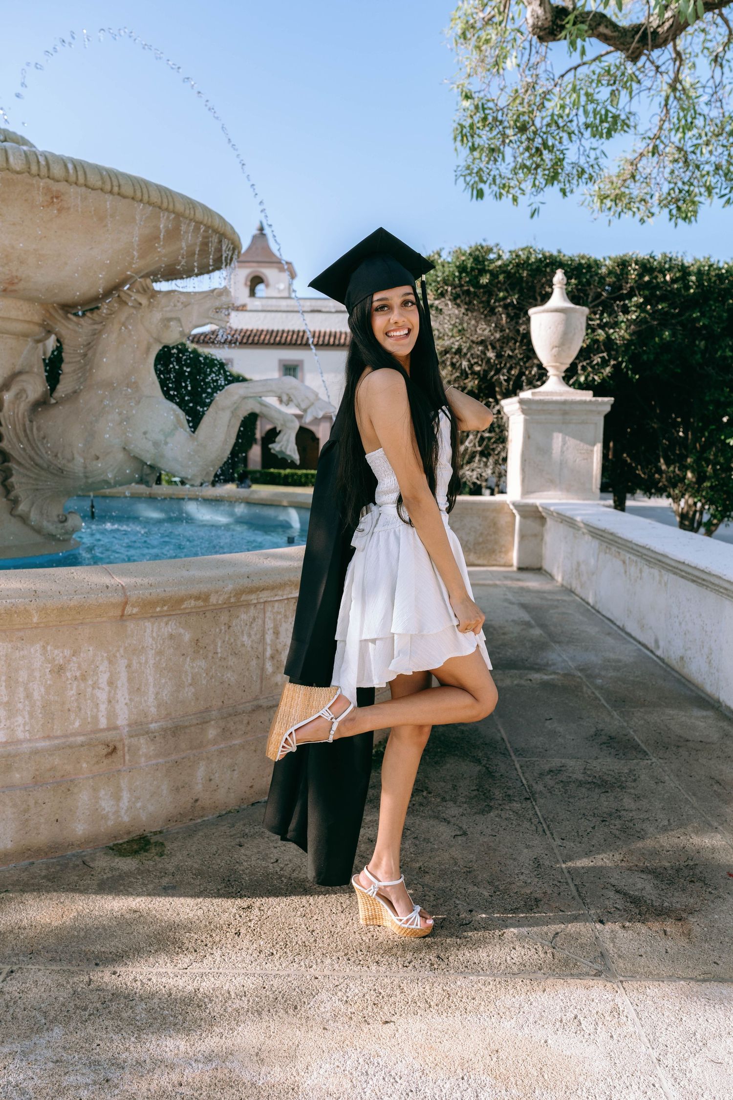 Smiling graduate in cap and gown posing by stone fountain outdoors on Worth Avenue Palm beach.
