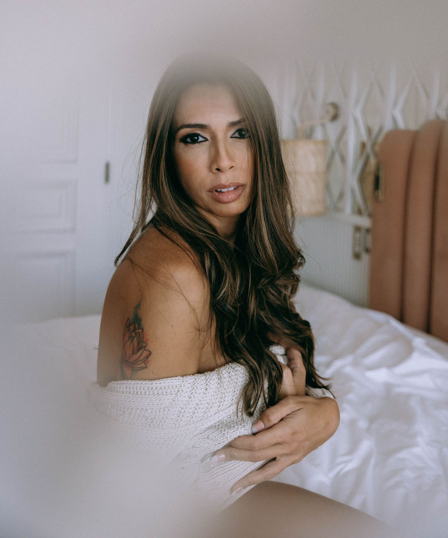 A young woman with long wavy brunette hair and a shoulder tattoo poses in a white off-shoulder wrap in a bright bedroom.  Boudoir Palm Beach
