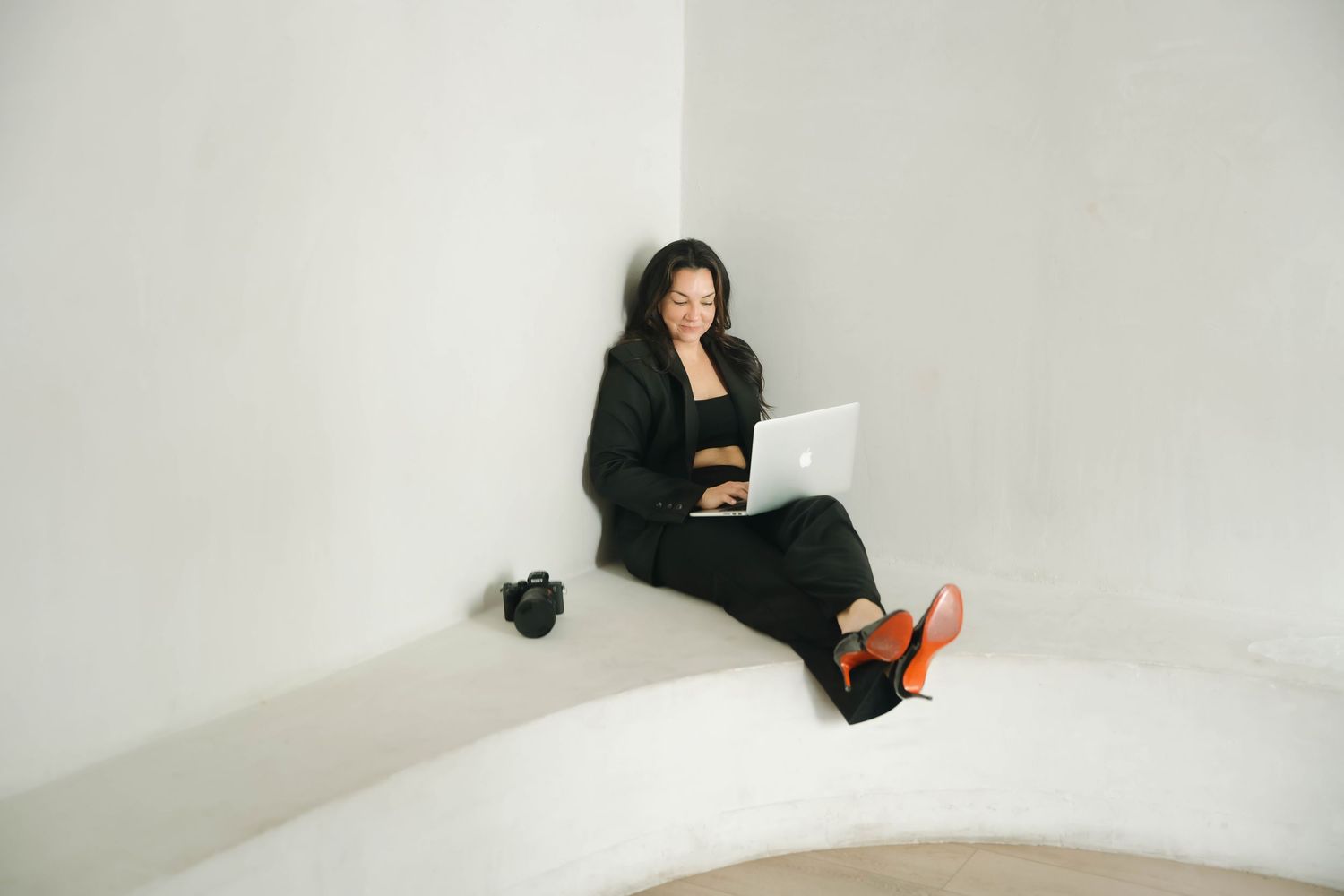 Person in black clothing sits reading against white walls in a minimalist interior setting.