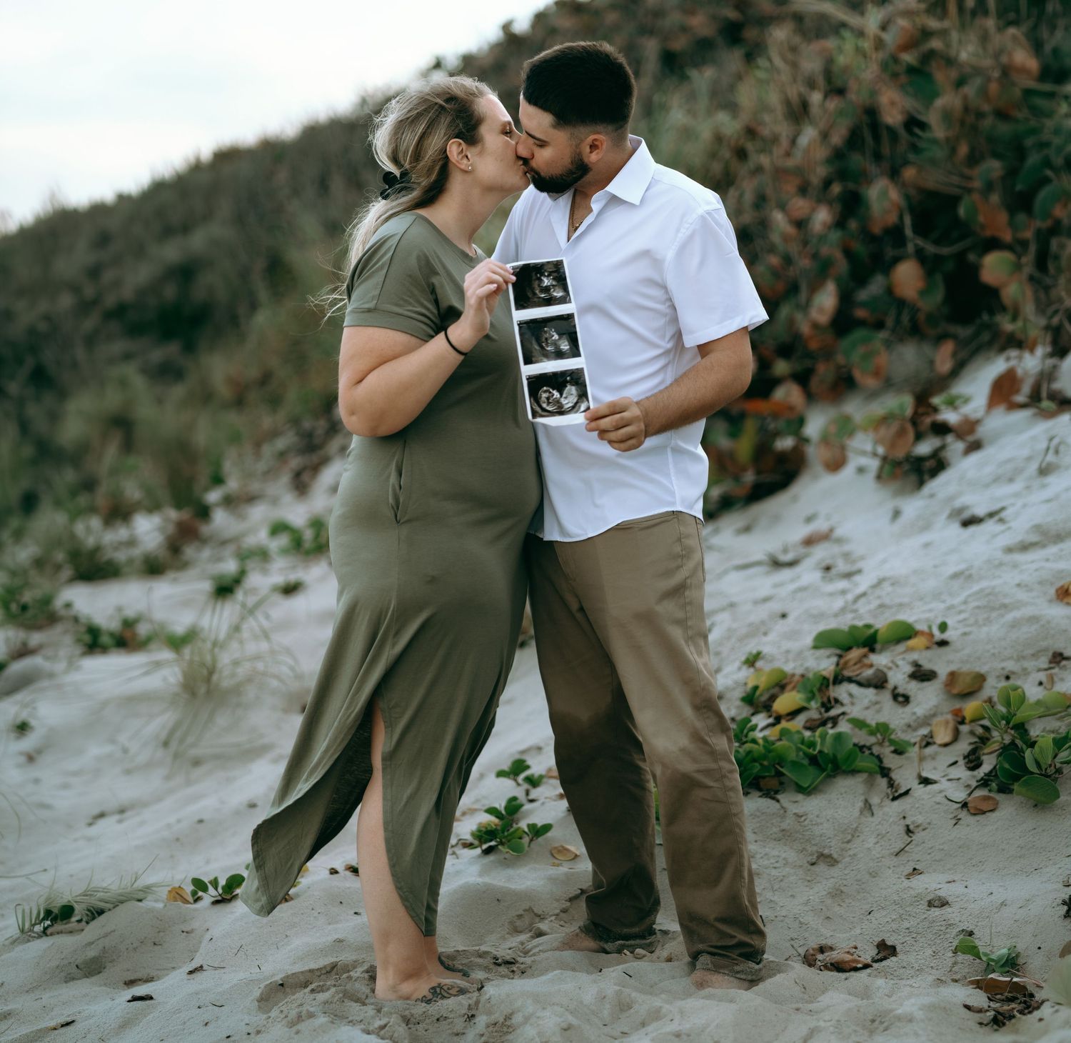 A couple shares tender moments on a sandy beach during a pregnancy announcement photoshoot.