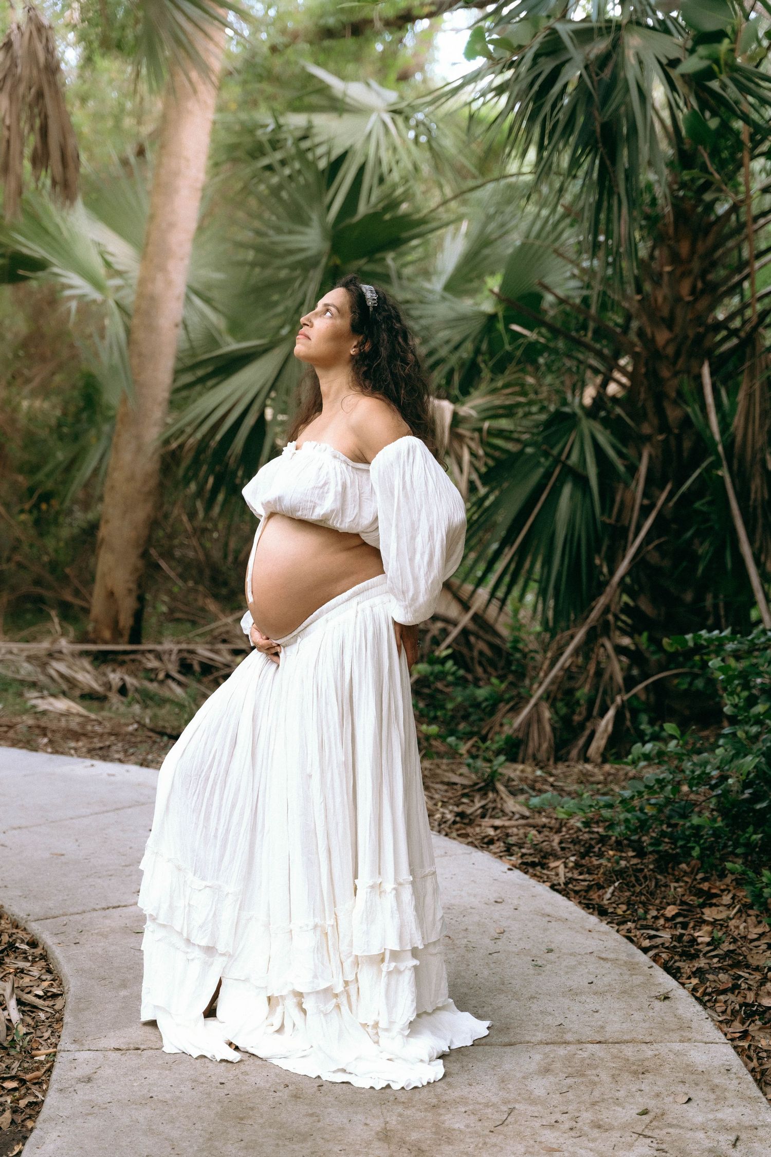 A woman in a flowing white dress poses gracefully on a stone path surrounded by tropical foliage.