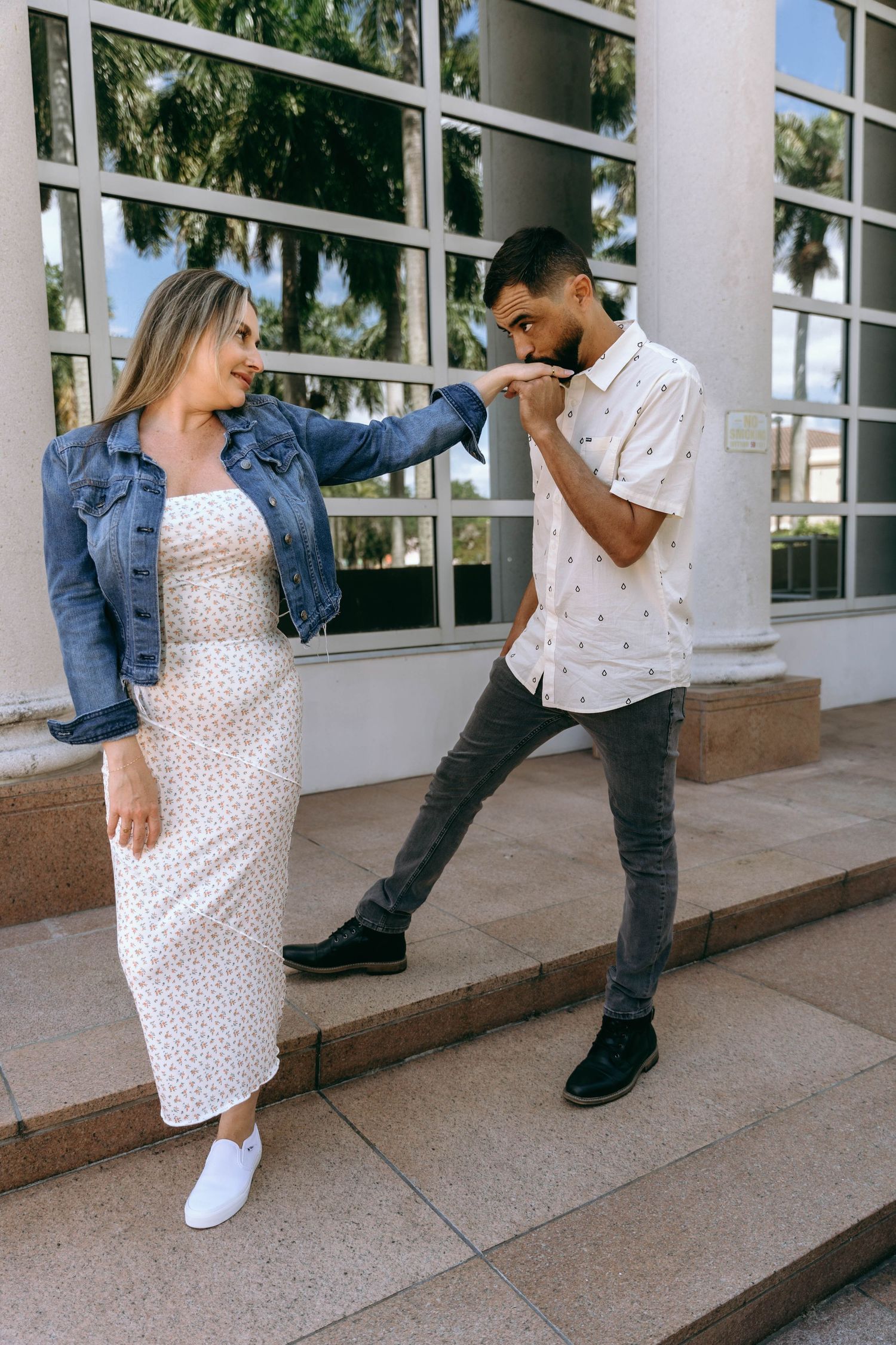 Playful couple poses on outdoor steps near glass windows in casual outfits.