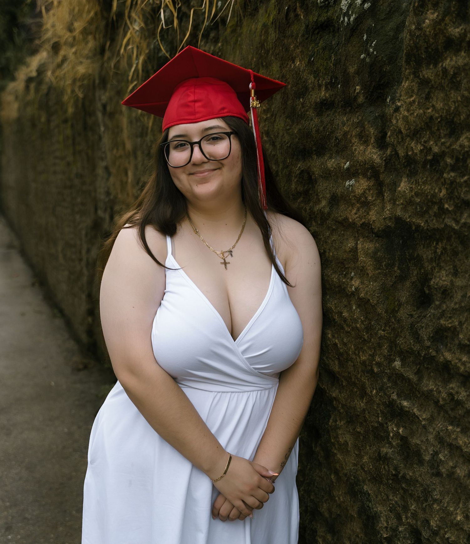 A graduate in a white dress and red cap poses against a stone wall.