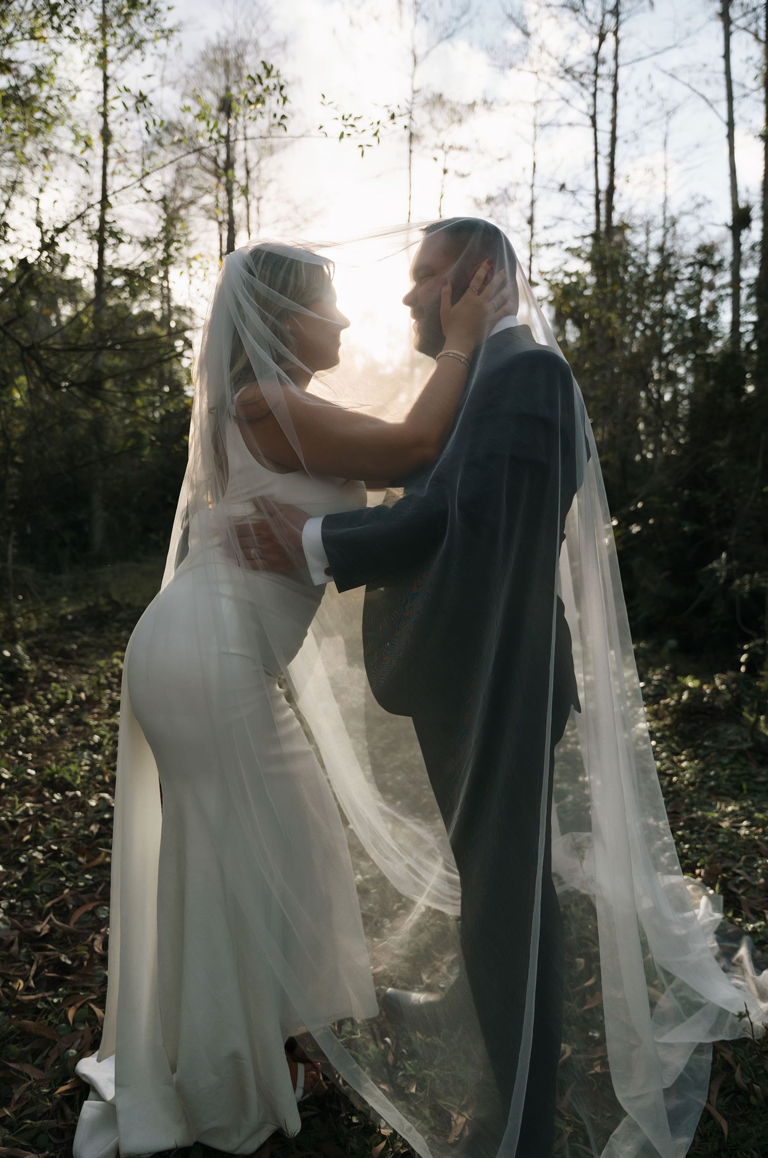 Sunlit wedding couple dancing in forest with flowing veil creating ethereal effect.