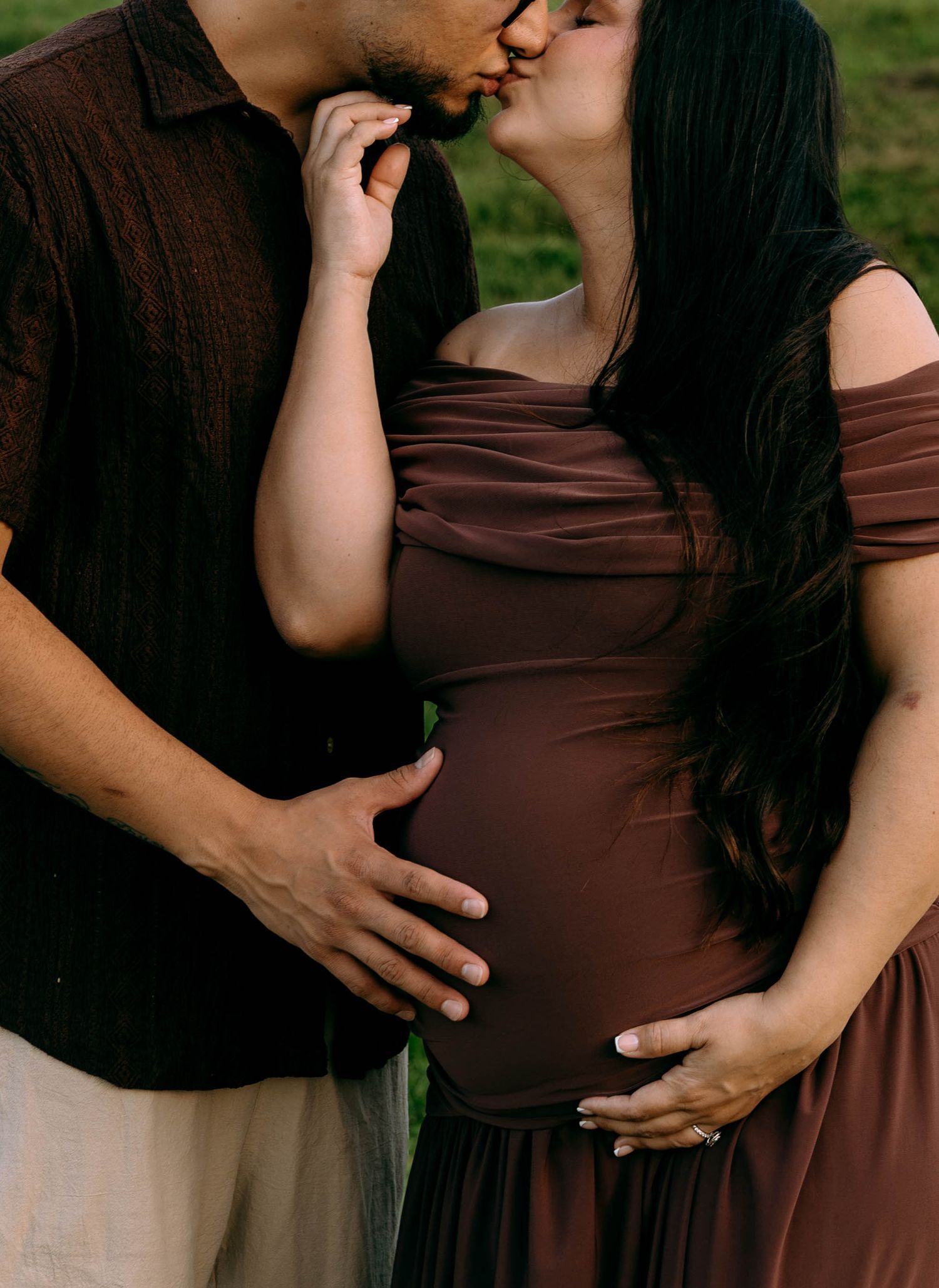 Couple sharing a tender kiss during a maternity photo shoot, woman in brown off-shoulder dress cradling baby bump outdoors.