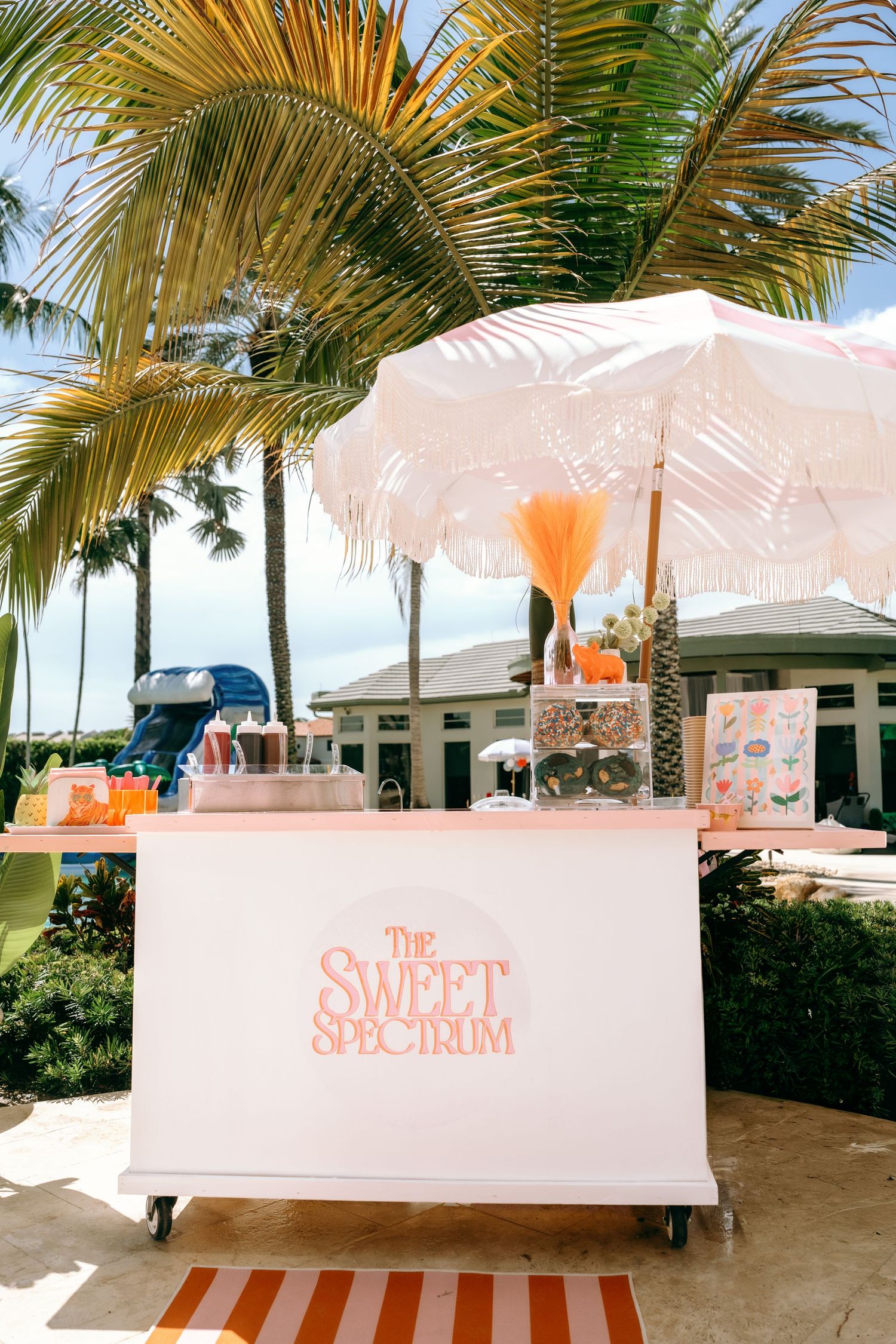 The Sweet Spectrum candy cart with white umbrella and palm trees at an outdoor tropical event.