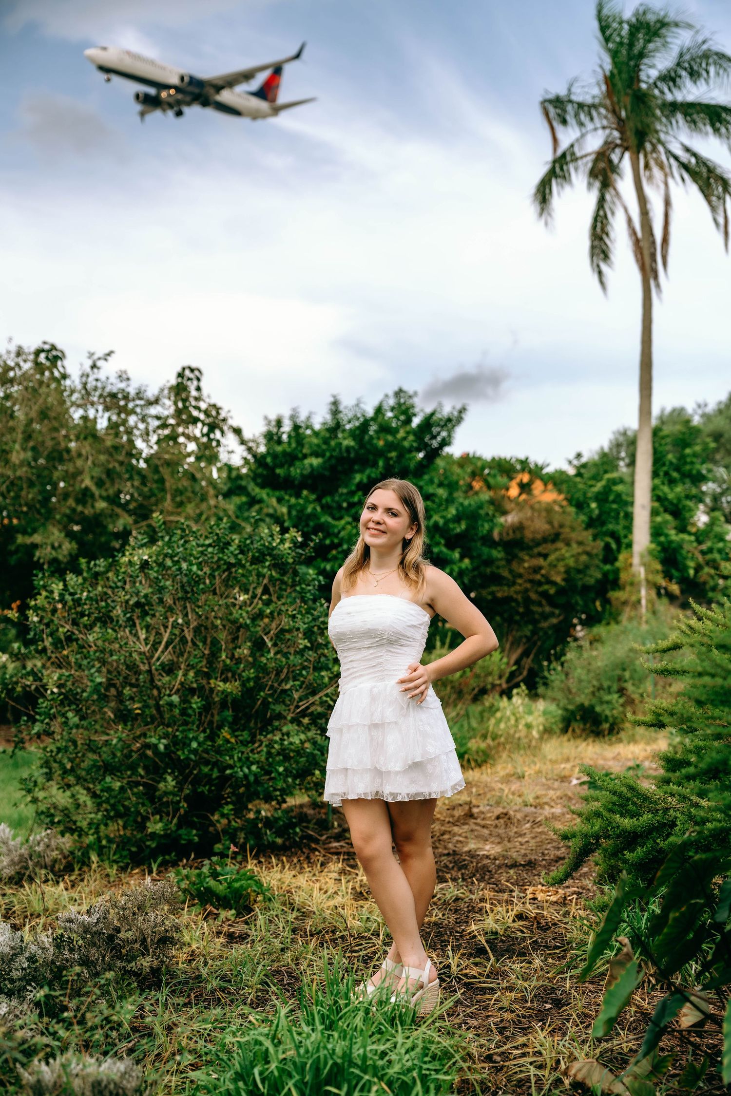 Woman in white dress stands in garden with Delta airplane flying overhead near palm tree.