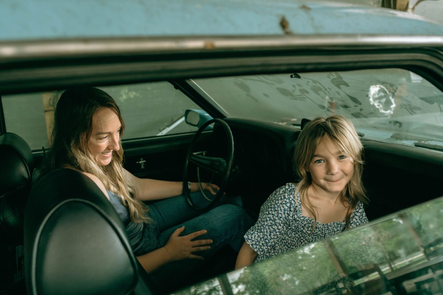 Two smiling girls sitting inside a vintage car, enjoying a fun moment together.