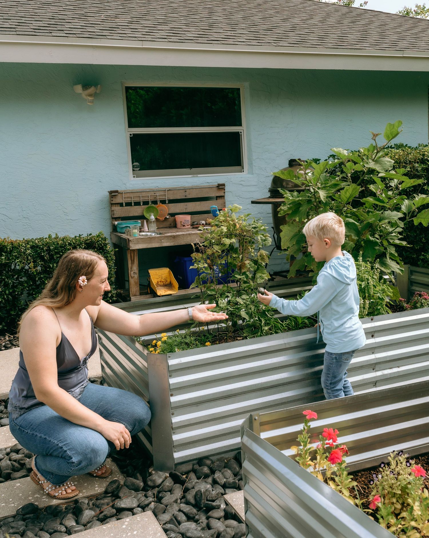 Woman and child interact over a raised metal garden bed outside a light blue house with lush greenery.