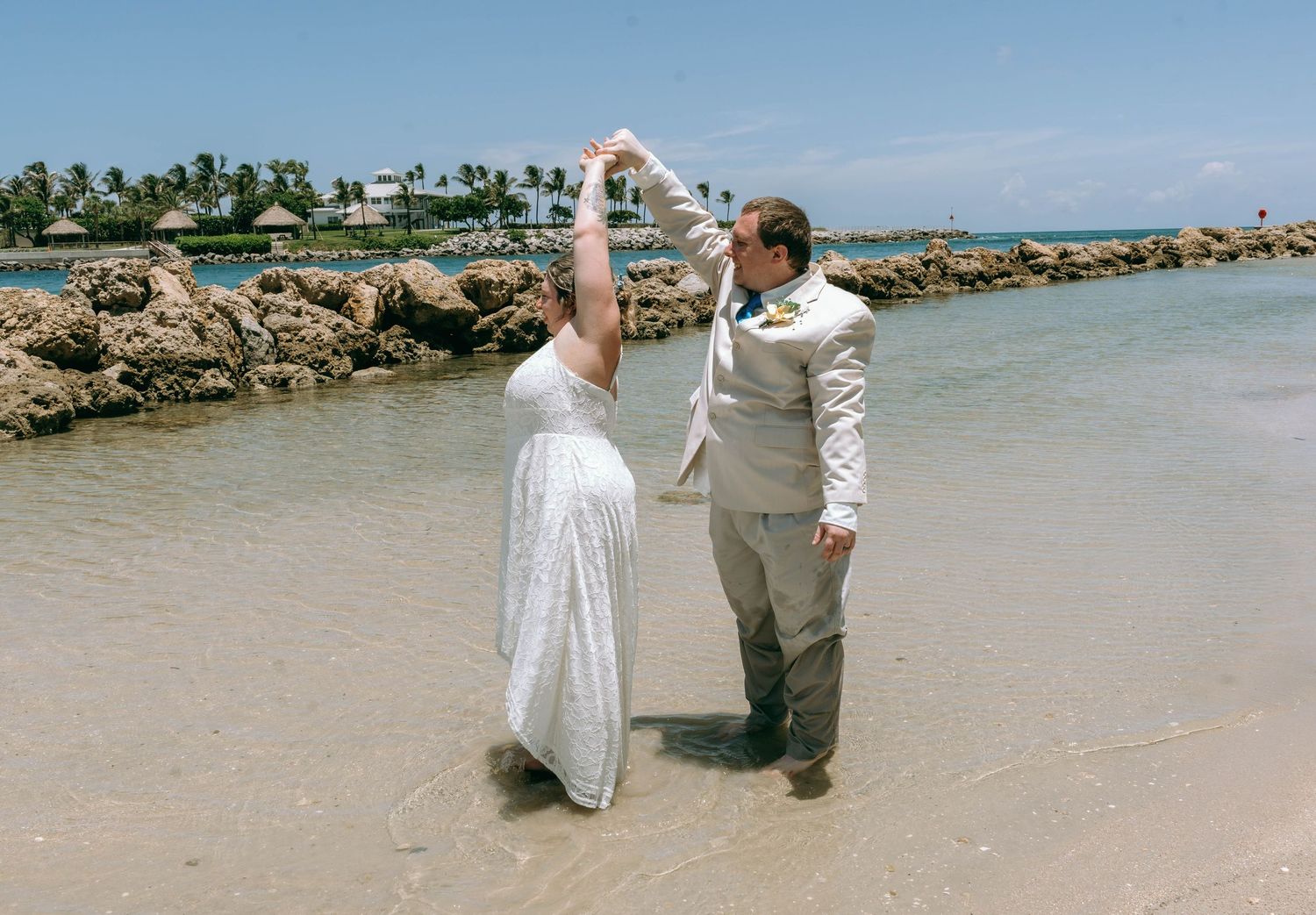 Bride and groom dancing on a tropical beach shoreline with rocky coastline and palm trees in the background.
