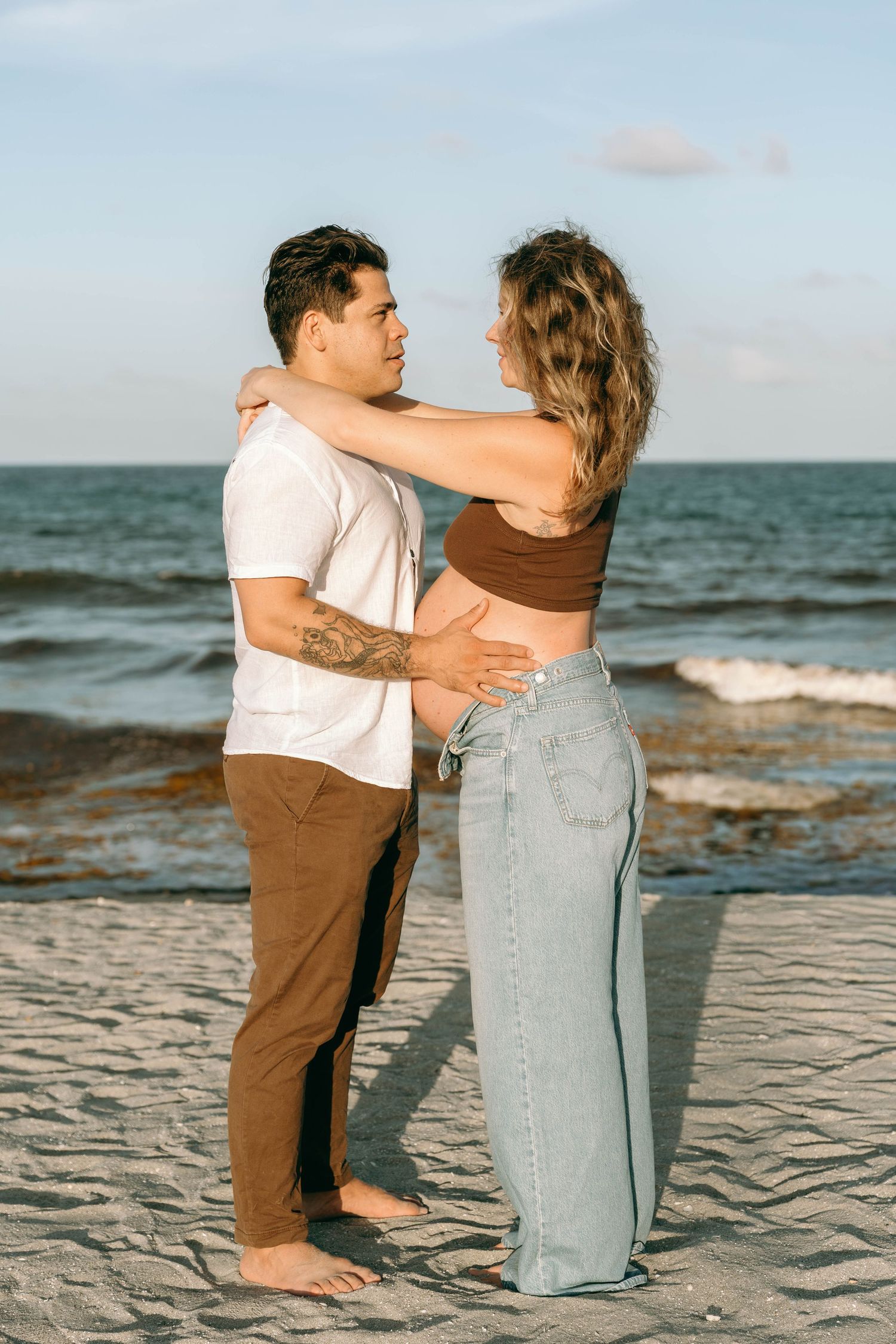 A couple embraces romantically on a sandy beach at sunset, with ocean waves in the background.
