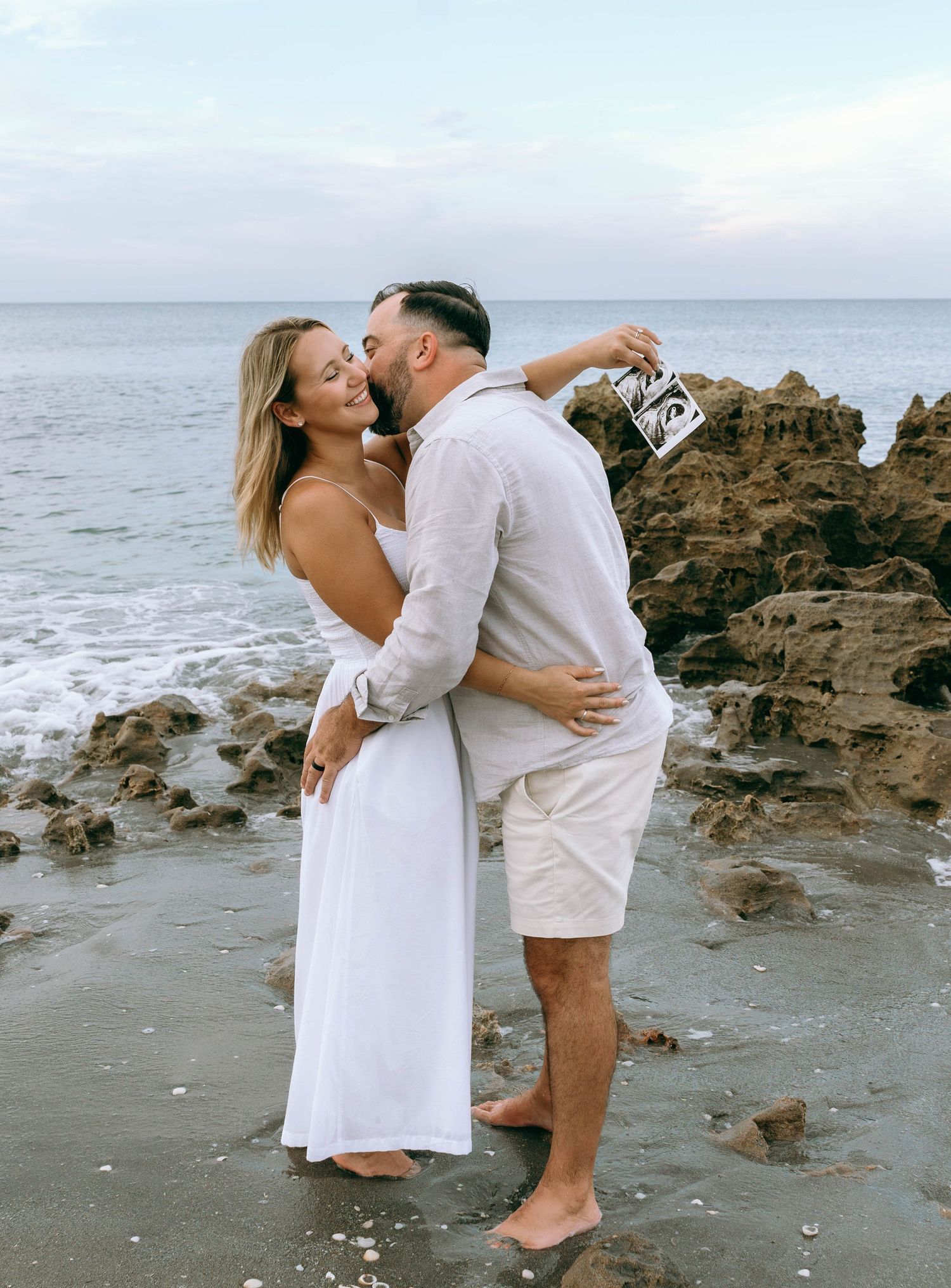 Couple embracing on rocky beach at sunset, woman in white dress, man in white shirt and shorts, holding ultrasound photo.
