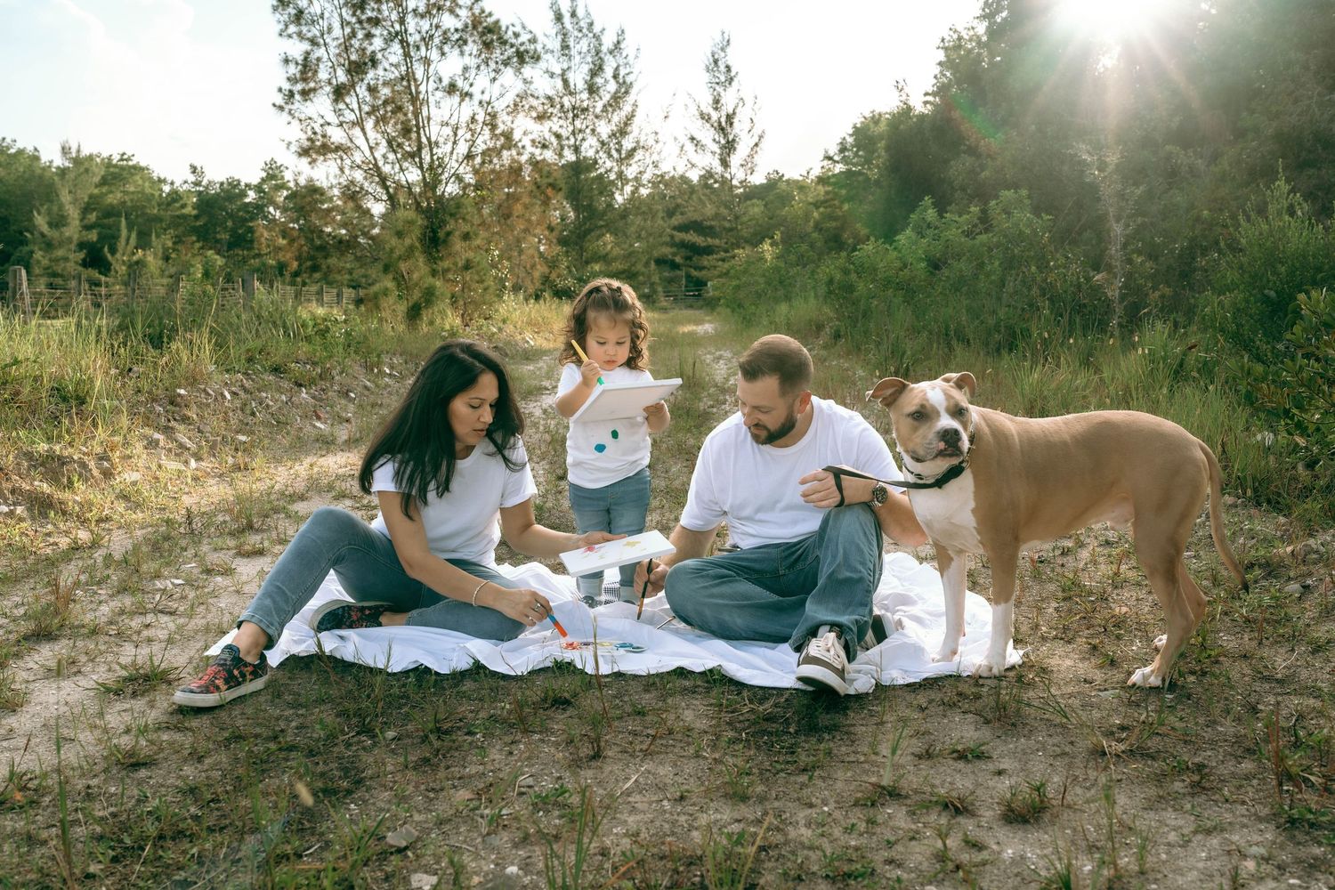 Family with young child and dog enjoying a sunny outdoor picnic on a blanket in a natural setting.