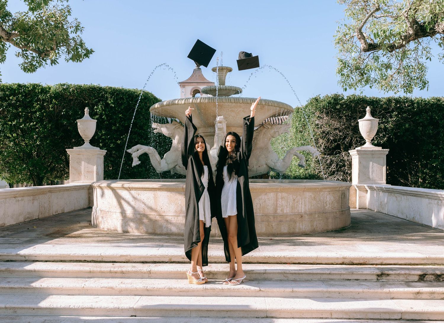 Two graduates in black robes toss caps by a fountain, celebrating their academic achievement outdoors on Worth Avenue Palm beach.