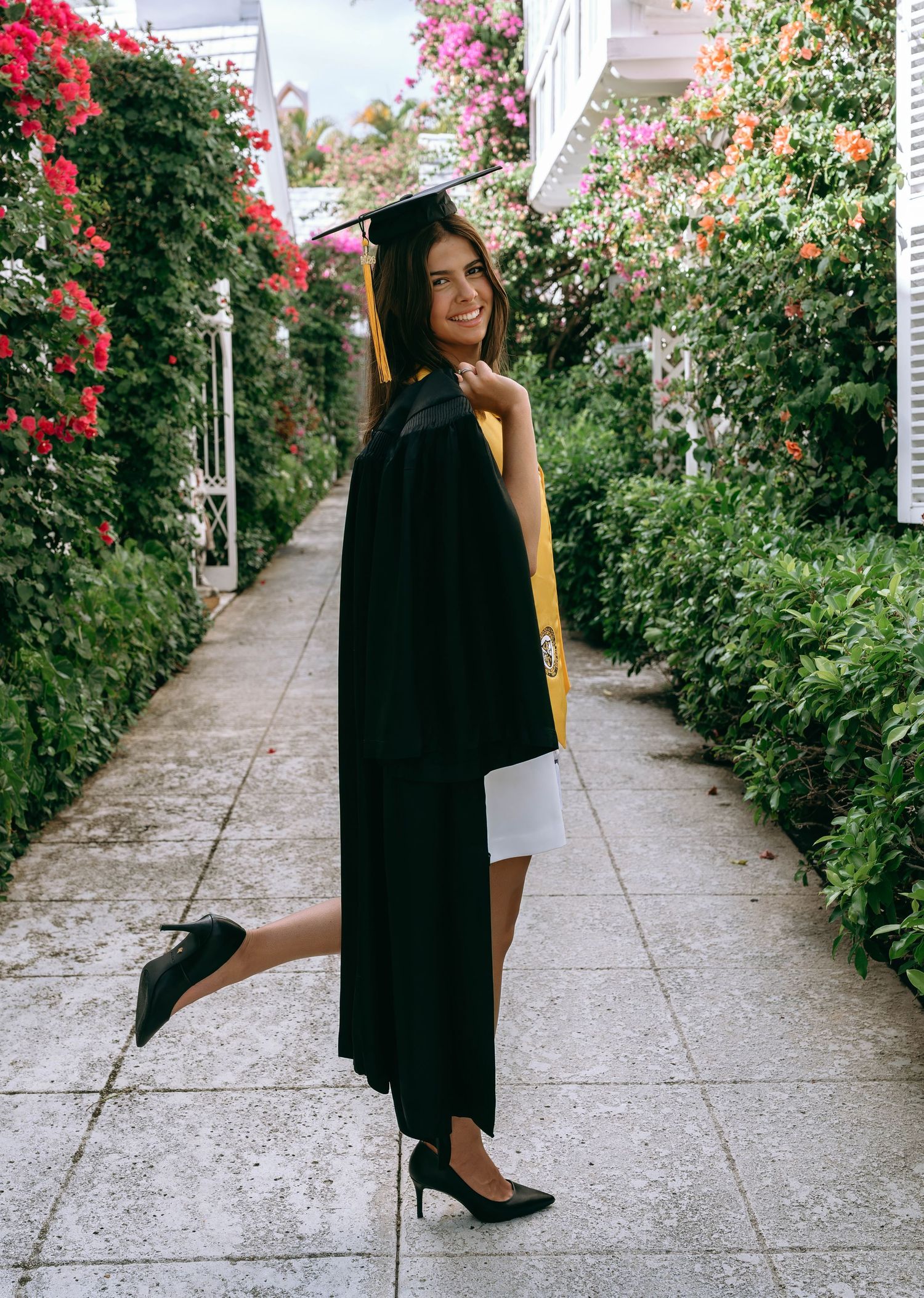 Graduate in black cap and gown poses playfully on flower-lined garden pathway in heels on Worth Avenue Palm beach.