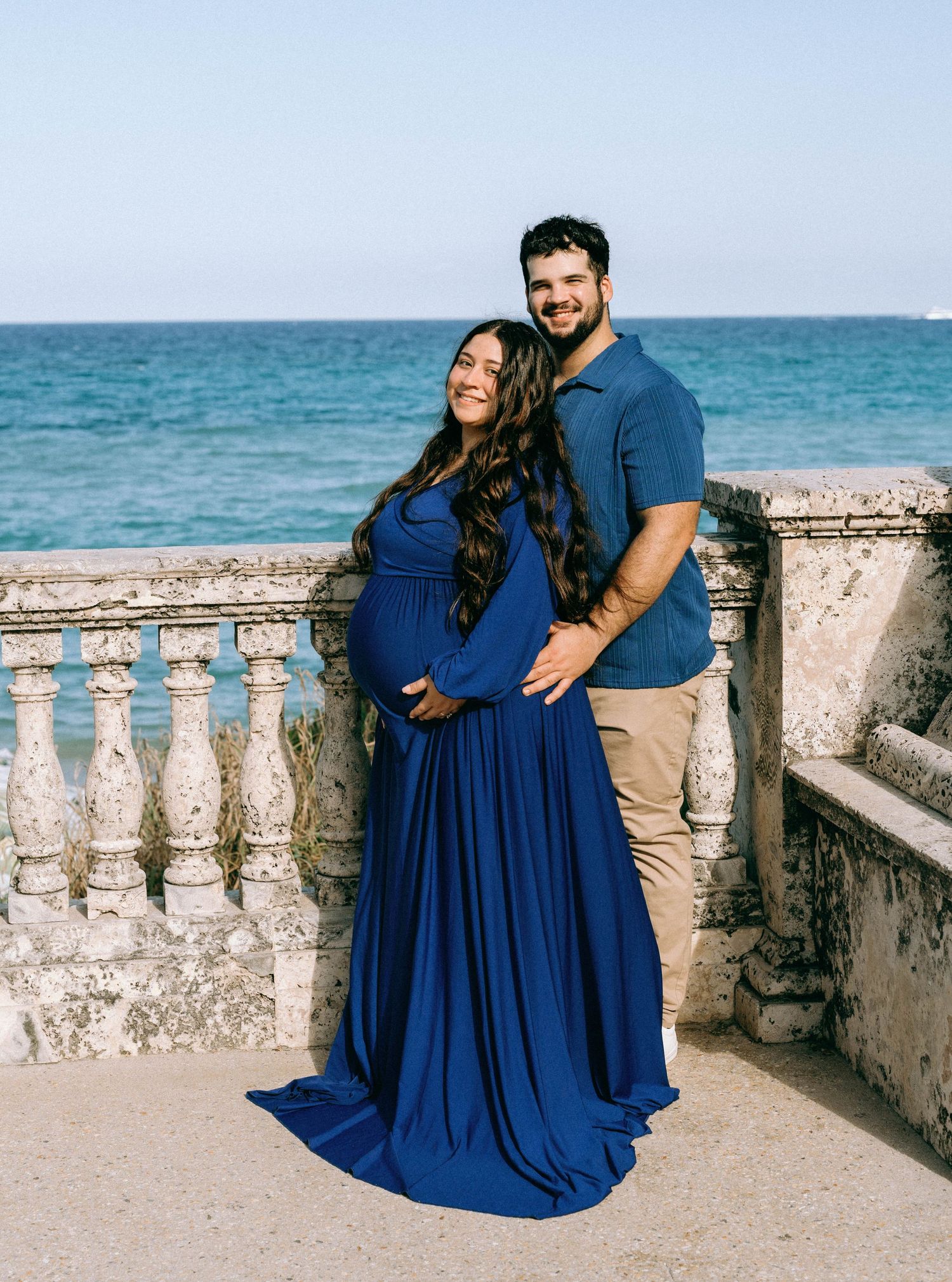 Pregnant woman in royal blue maternity gown poses with partner on stone balcony overlooking the ocean. Worth Avenue, Palm Beach, FL