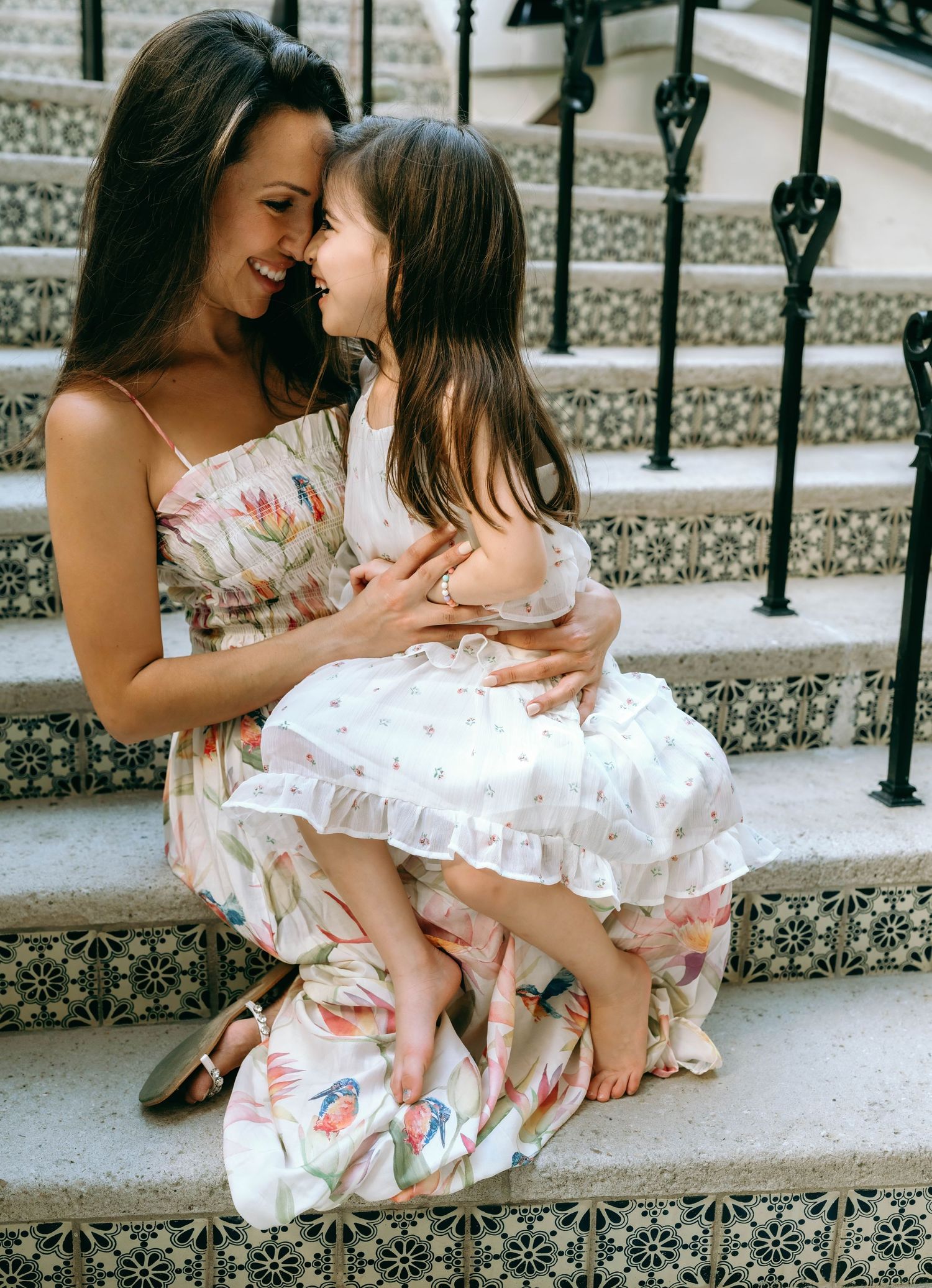Smiling mother in floral dress sits on tiled steps holding young daughter in white ruffled dress. Worth Avenue, Palm Beach, FL