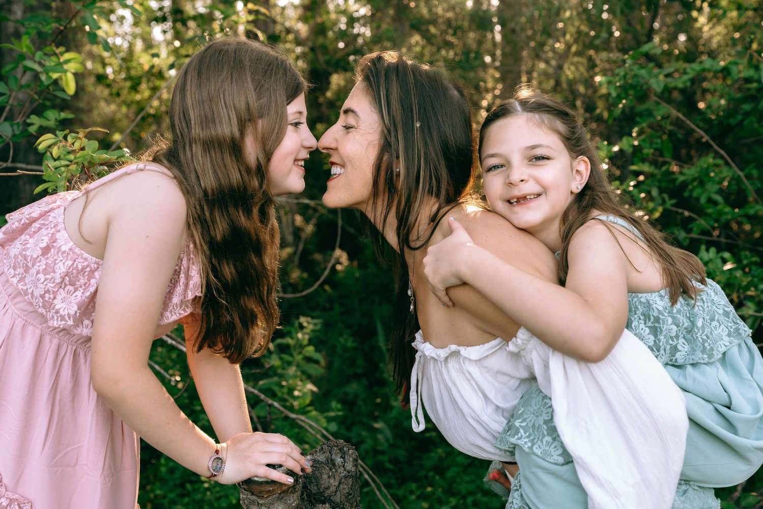 Two girls in pink and white dresses laugh and hug outdoors surrounded by lush green foliage. Colt Farms