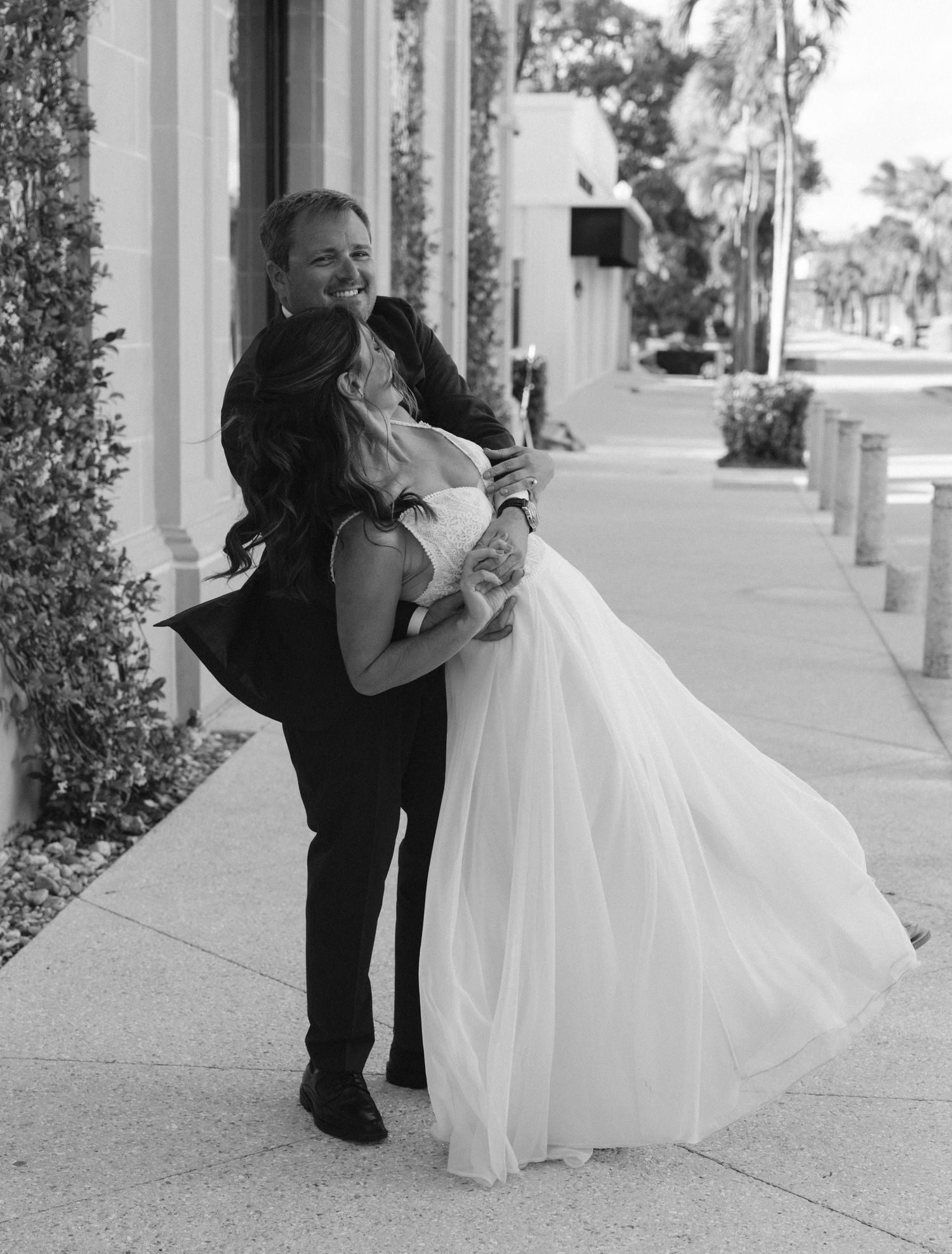 Black and white photo of groom lifting bride in flowing white dress on a sidewalk outside a building.