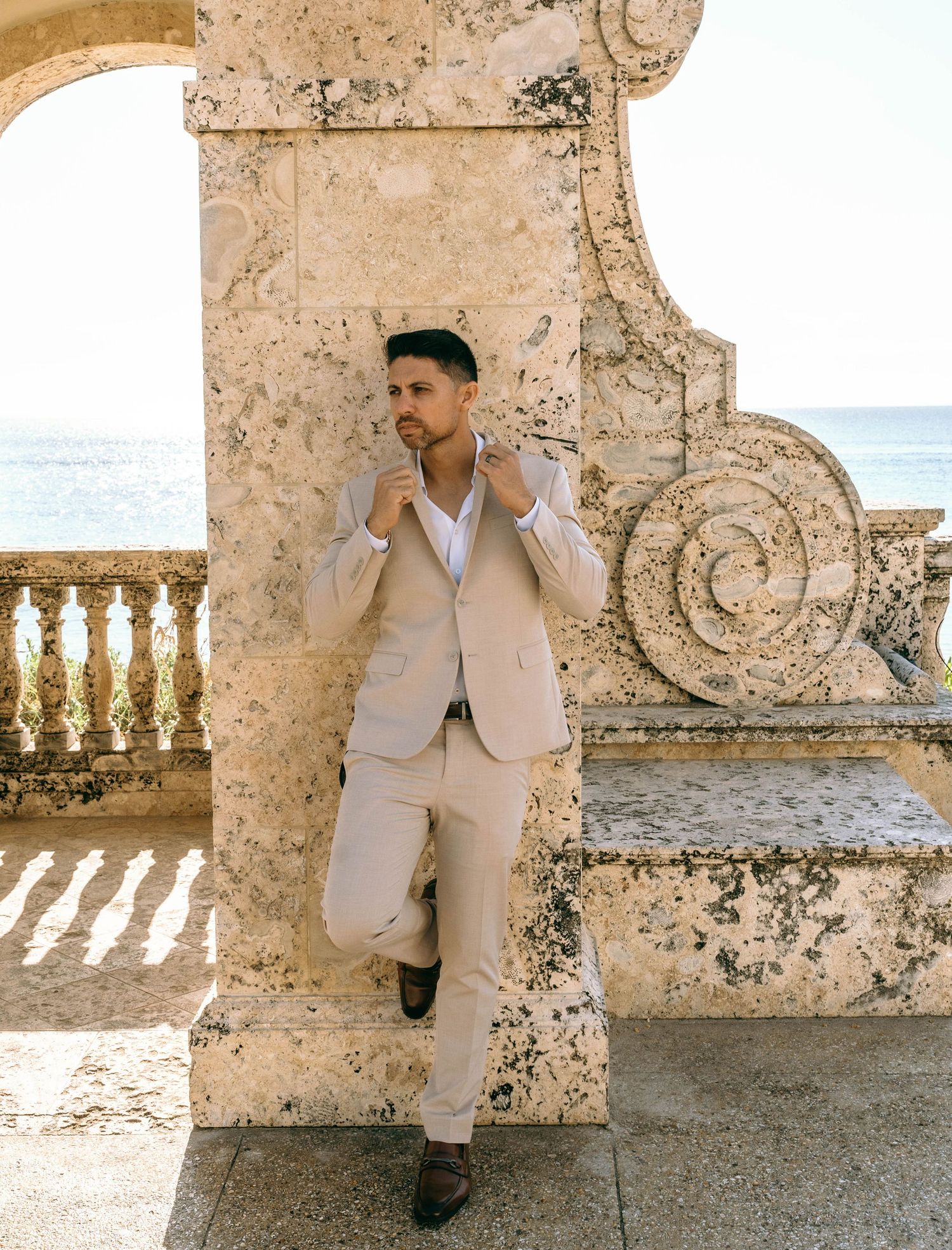 Groom in beige suit leaning against ornate stone column with ocean view backdrop.