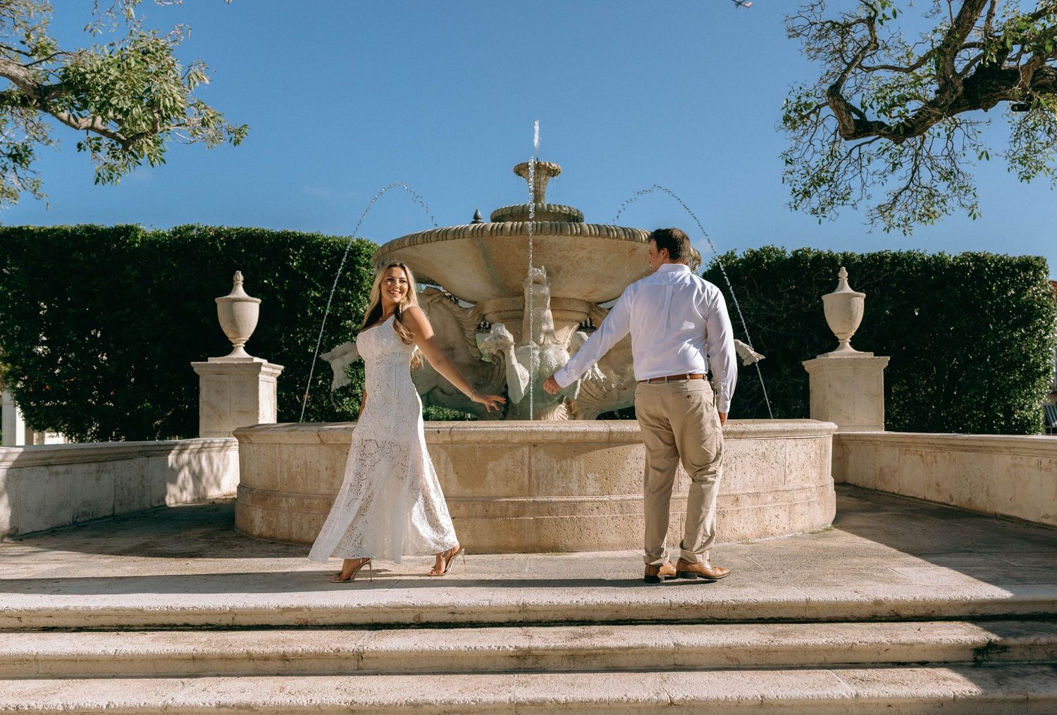 Couple dancing by ornate stone fountain in formal garden with manicured hedges on sunny day.