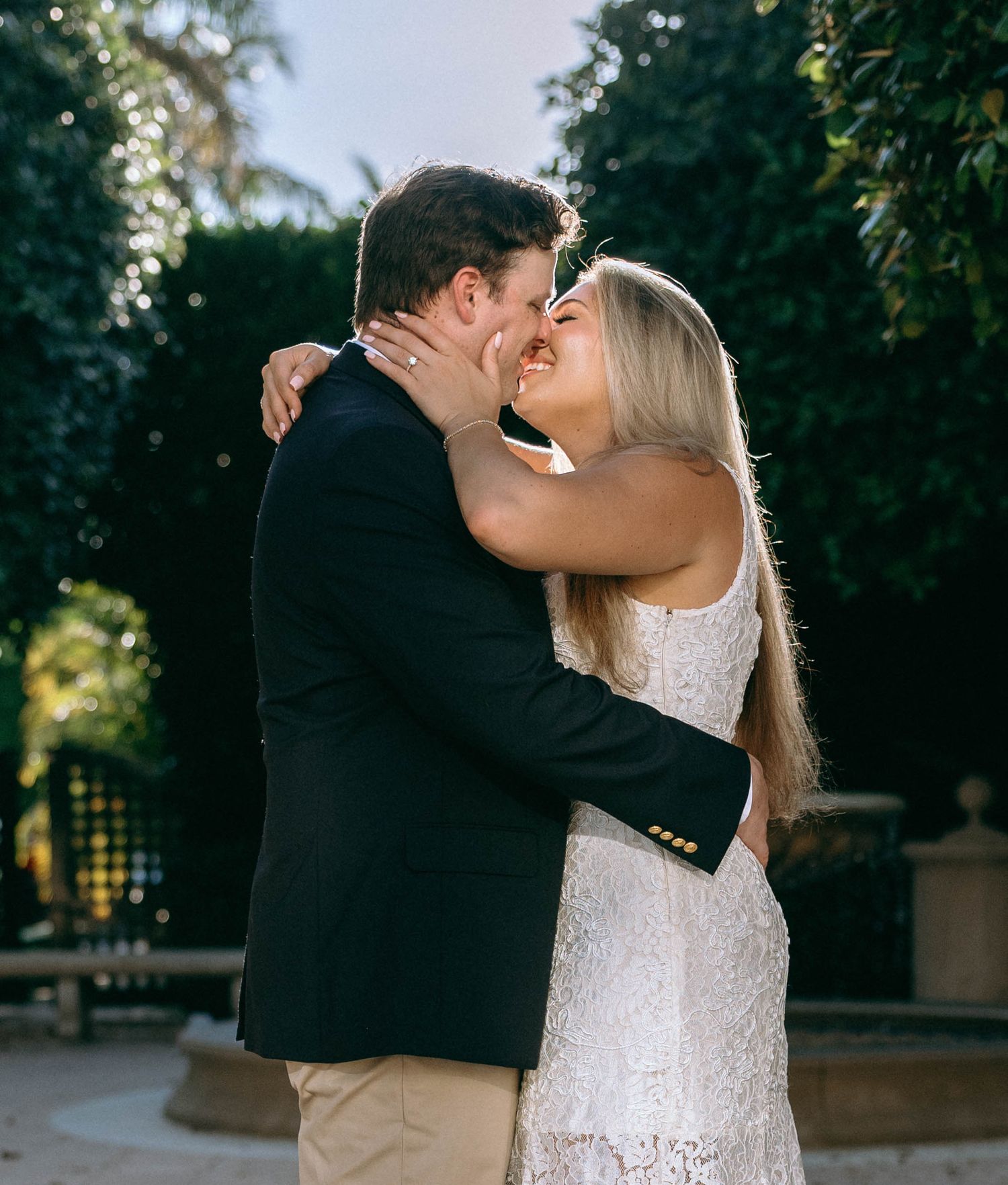Couple sharing a romantic kiss outdoors, woman in white lace dress embracing man in navy blazer near lush green hedges.