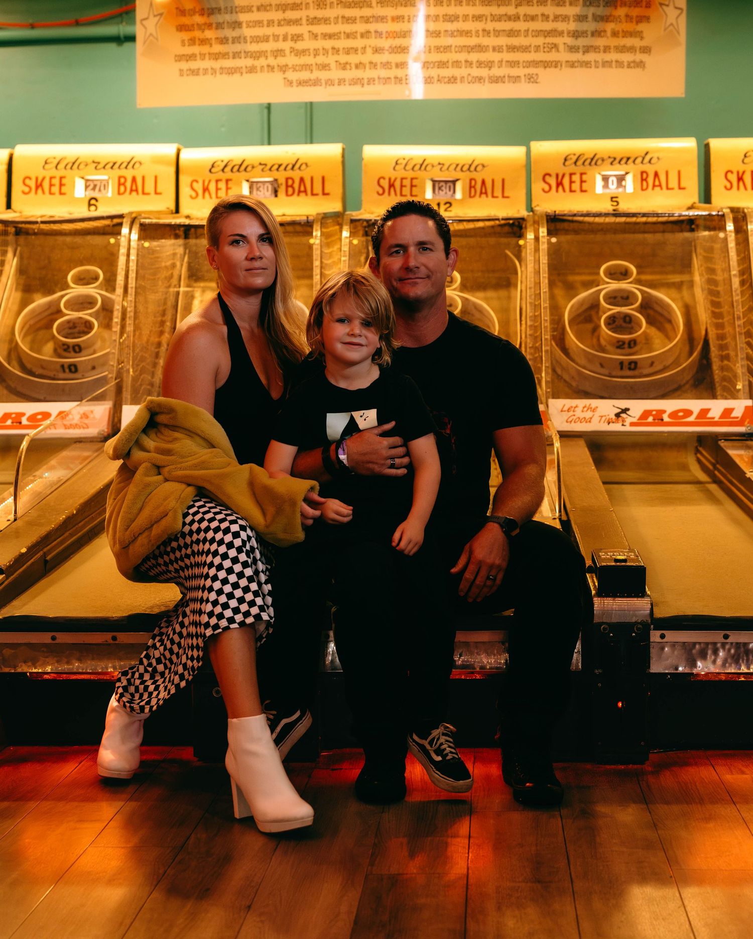 Family of three posing in front of Skee-Ball arcade games, parents in stylish outfits with young child between them.
