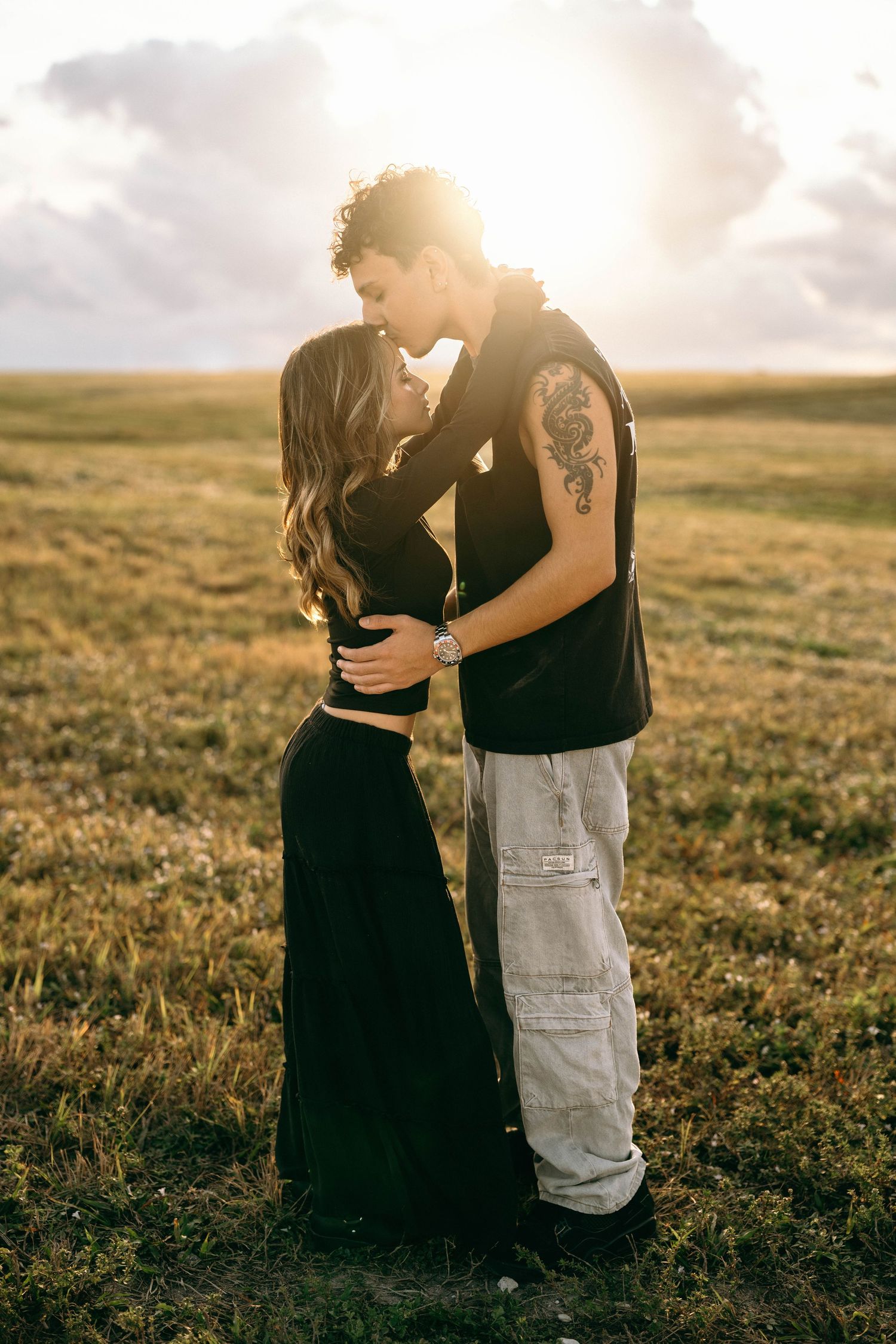 A romantic couple embraces in a golden sunset on a grassy field, with sun rays creating a dreamy backlit silhouette.