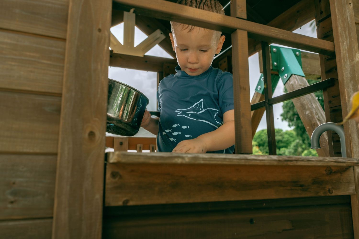 Young boy playing with a metal bucket in a wooden outdoor playhouse on a sunny day.