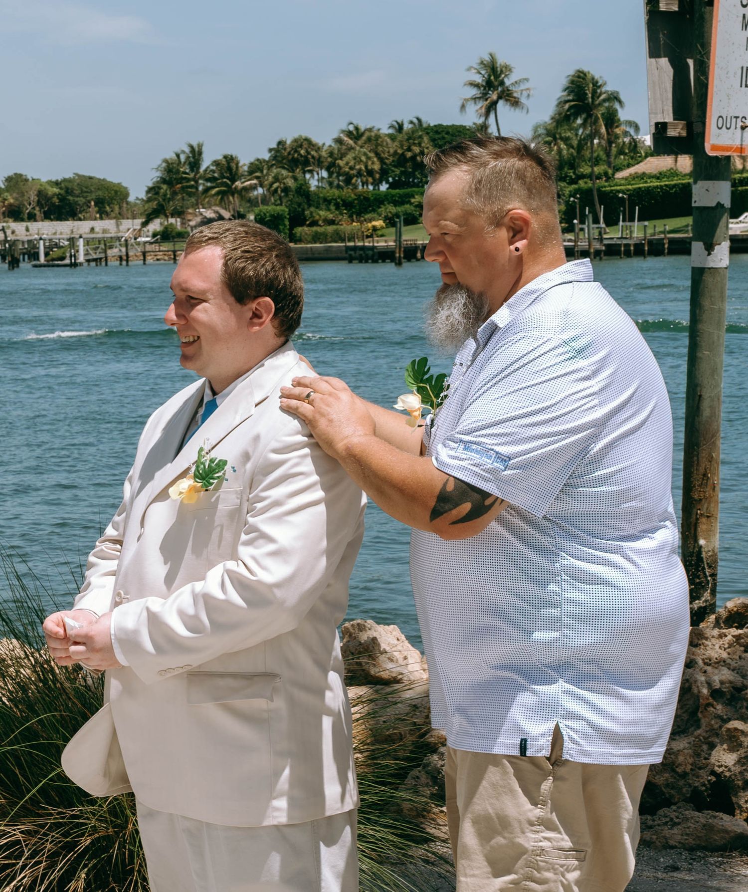 Groom in white suit gets boutonniere pinned by bearded man near tropical waterfront on sunny wedding day.