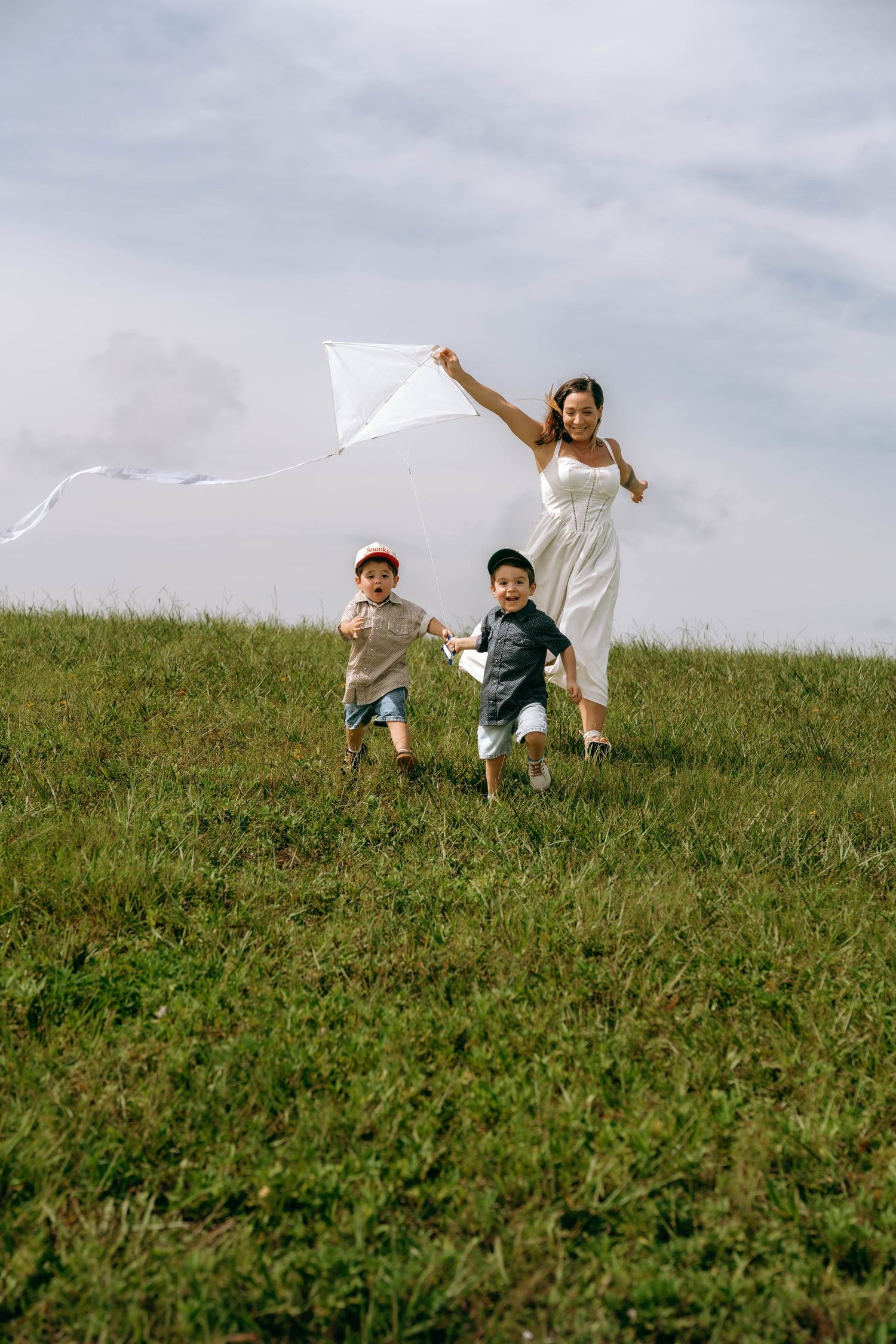 A joyful mother in a white dress runs with two young boys flying a white kite on a green grassy hill.