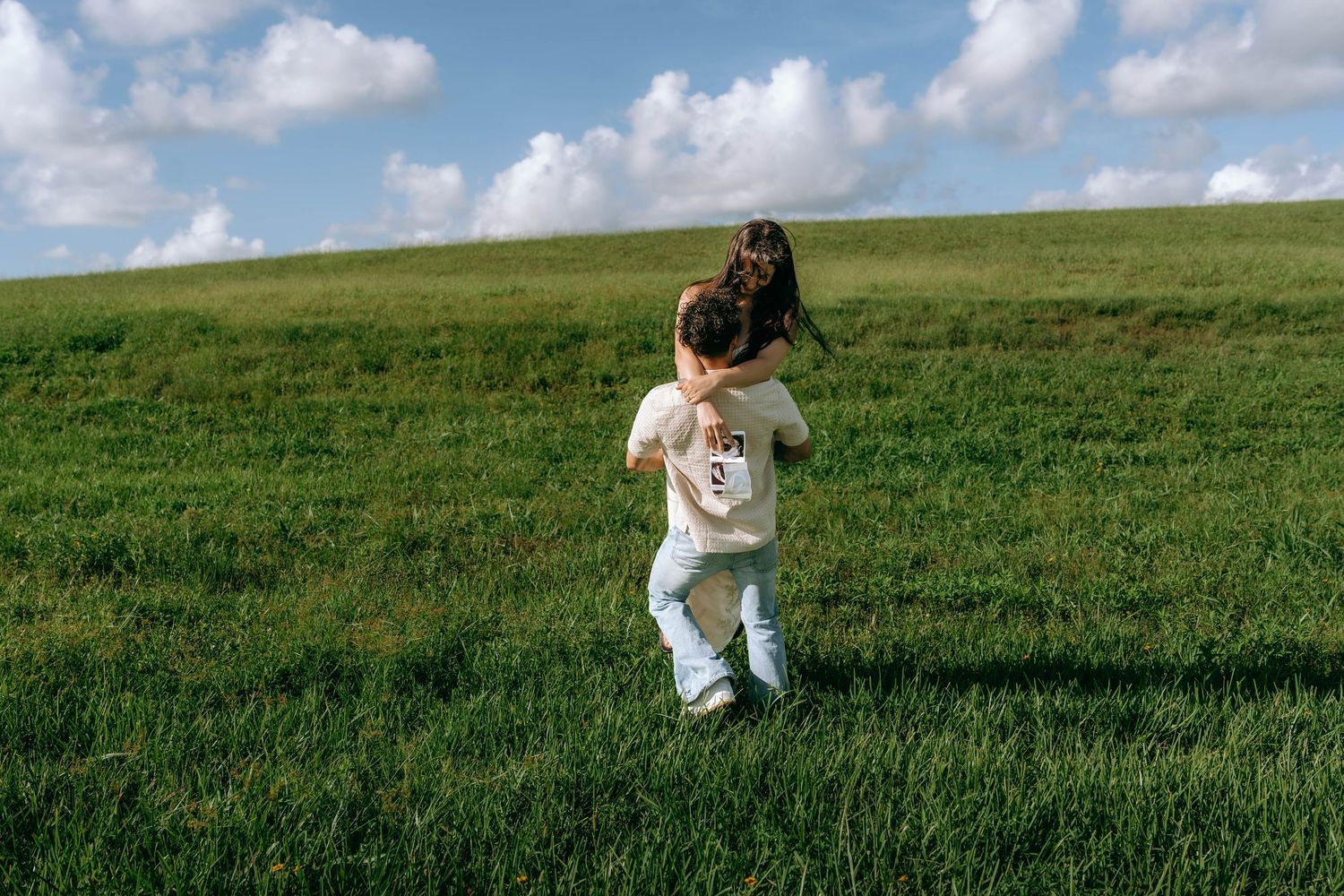 Two children playing and hugging on a lush green hillside under a bright blue sky with fluffy white clouds.
