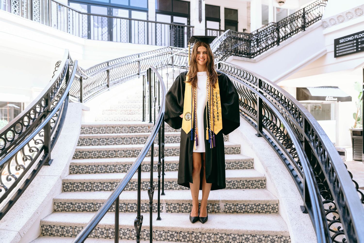 Graduate in black cap and gown standing on ornate staircase with gold honor cords on Worth Avenue Palm beach.