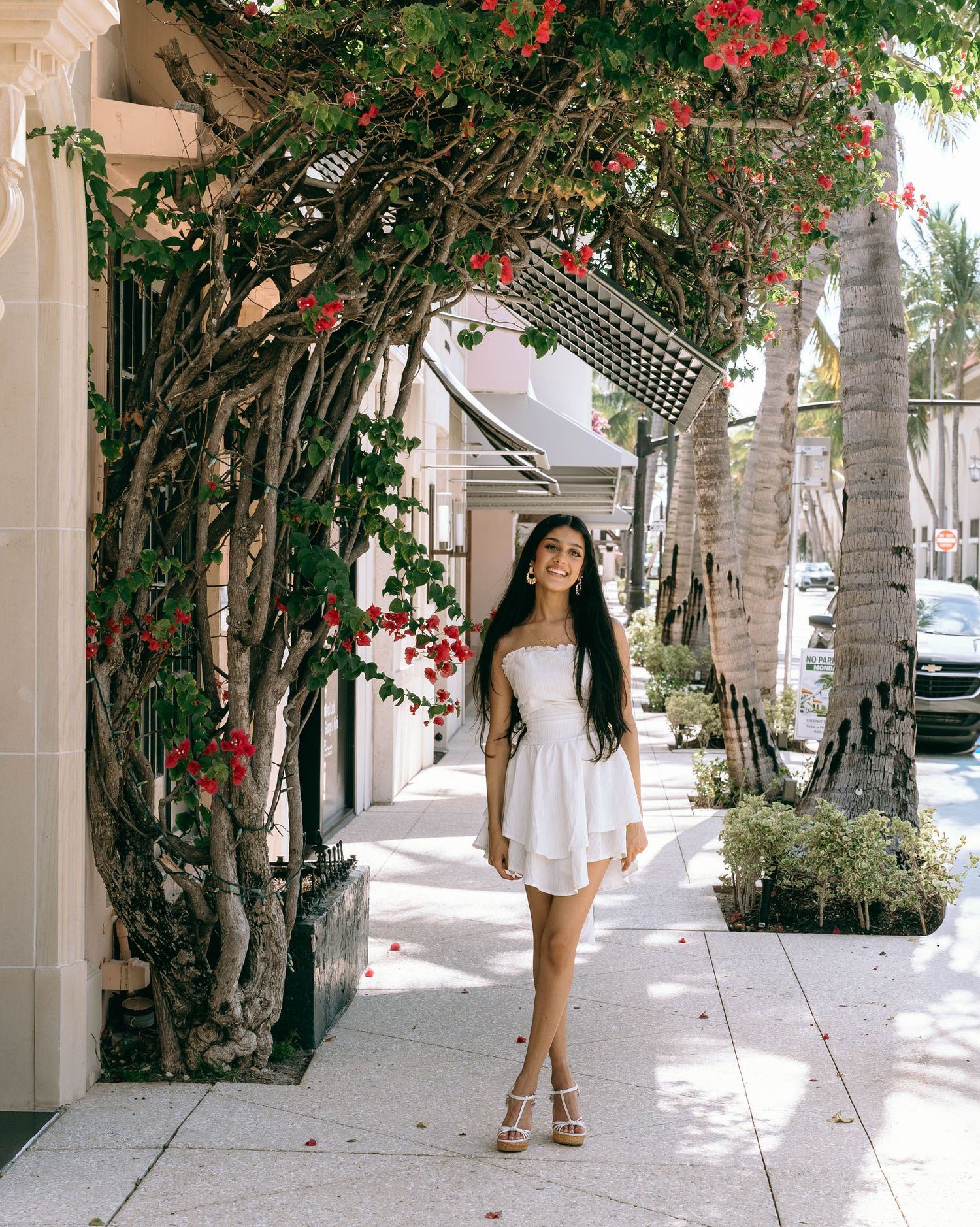 Young woman in white strapless dress posing under a bougainvillea arch on a sunny sidewalk on Worth Avenue Palm beach.
