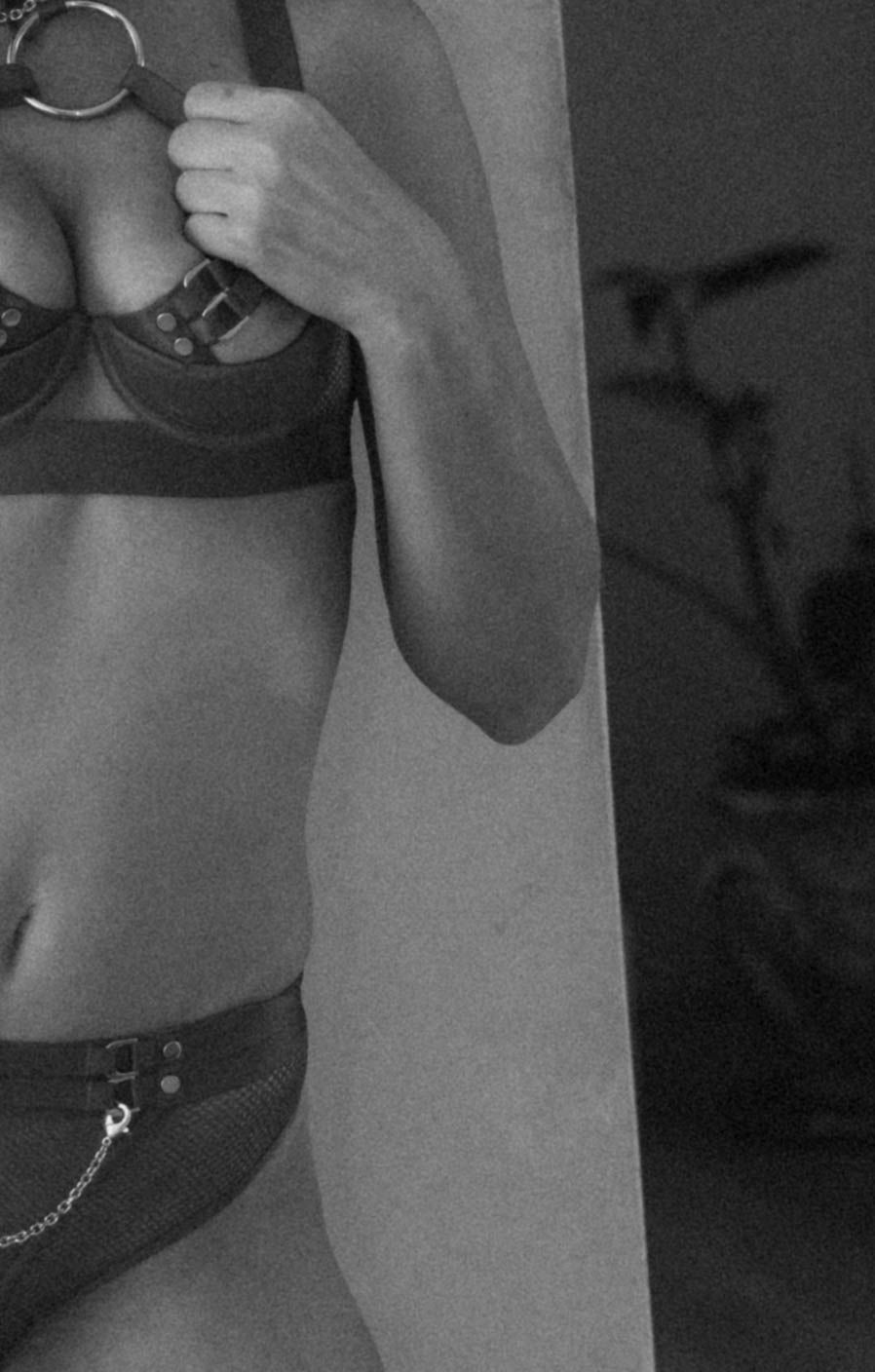 Black and white photo of a woman in a strappy bralette and chain-detail bikini bottoms posing indoors.